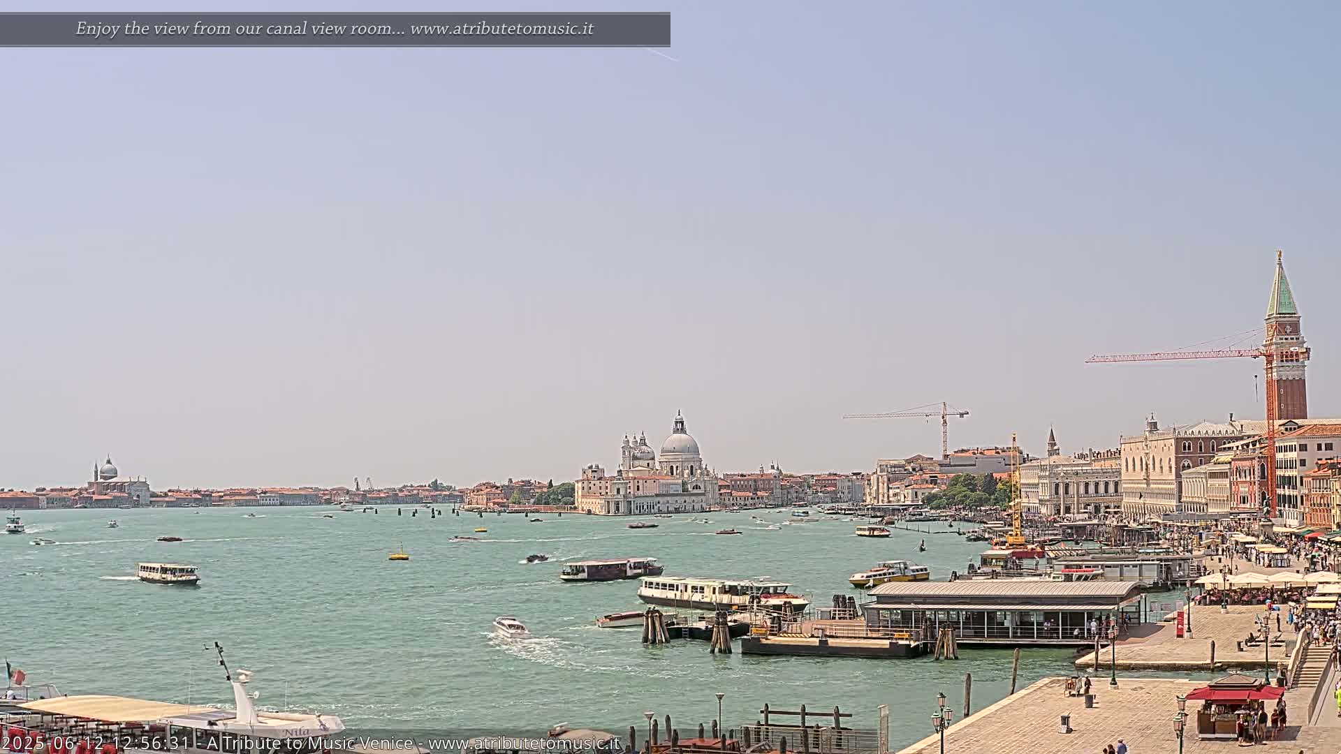 A sunny day in Venice shows a wide canal with numerous boats, a waterfront with buildings and people, and the iconic Basilica di Santa Maria della Salute in the distance.