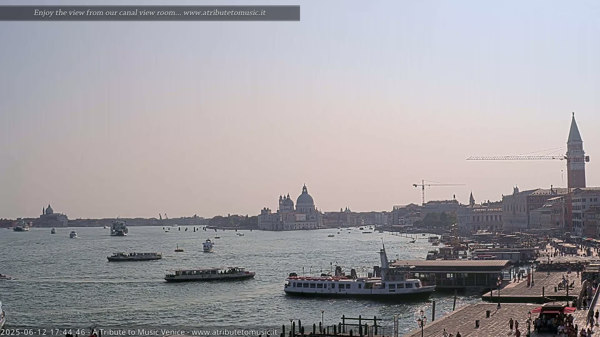 A hazy sunny day in Venice shows a wide canal with numerous boats, a cityscape featuring a domed church, and a tall bell tower.