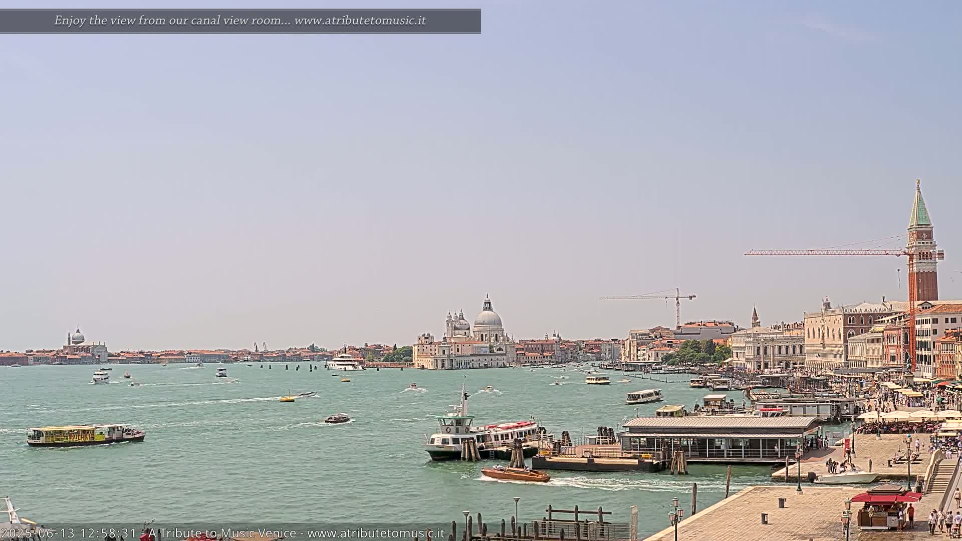 A sunny Venetian canal scene shows several boats, a ferry, and the Basilica di Santa Maria della Salute in the distance, with a construction crane visible along the waterfront.