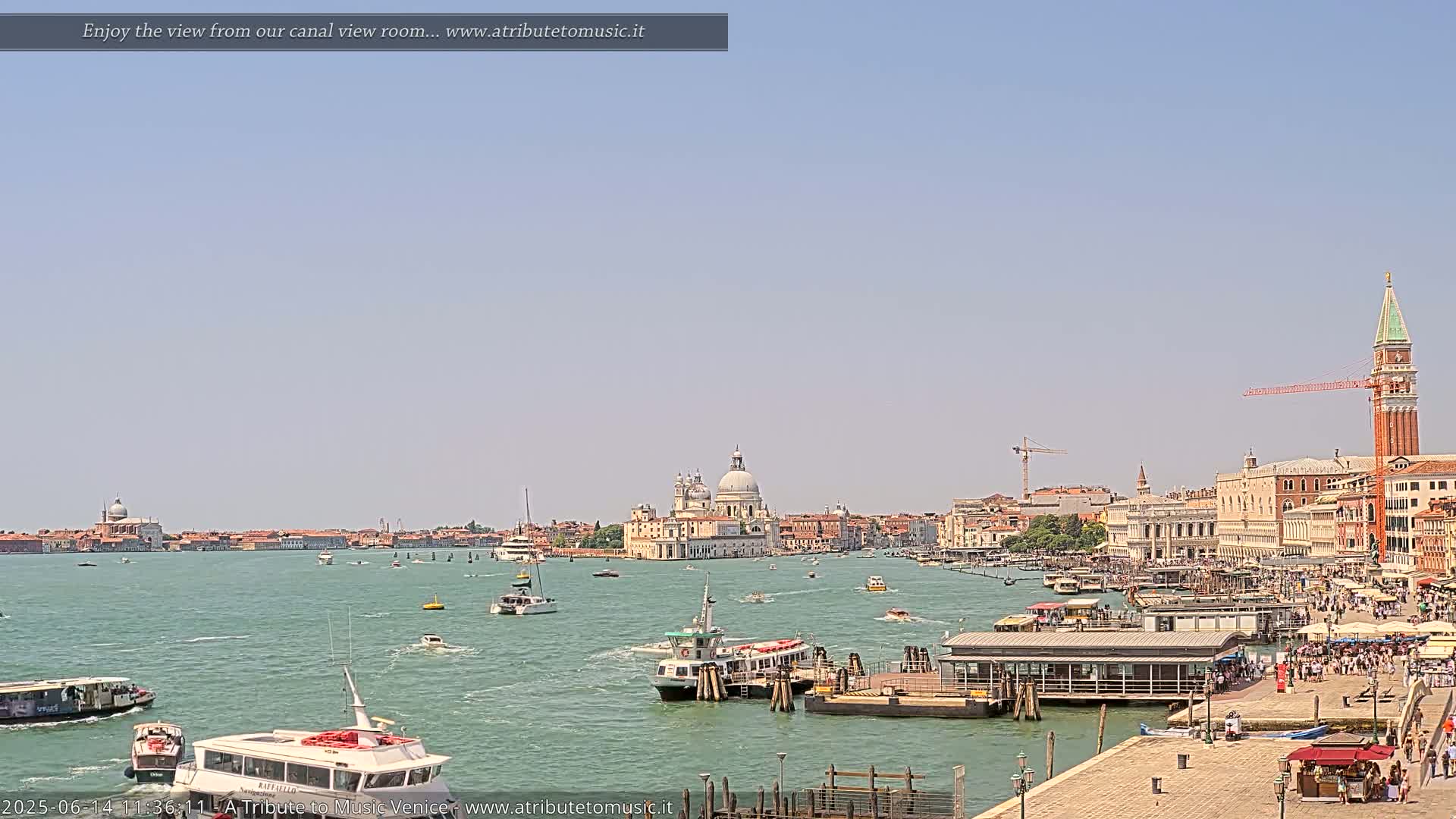 A sunny day in Venice shows a wide canal with numerous boats and a view of the city's buildings, including a large church with a dome, and a tall bell tower.