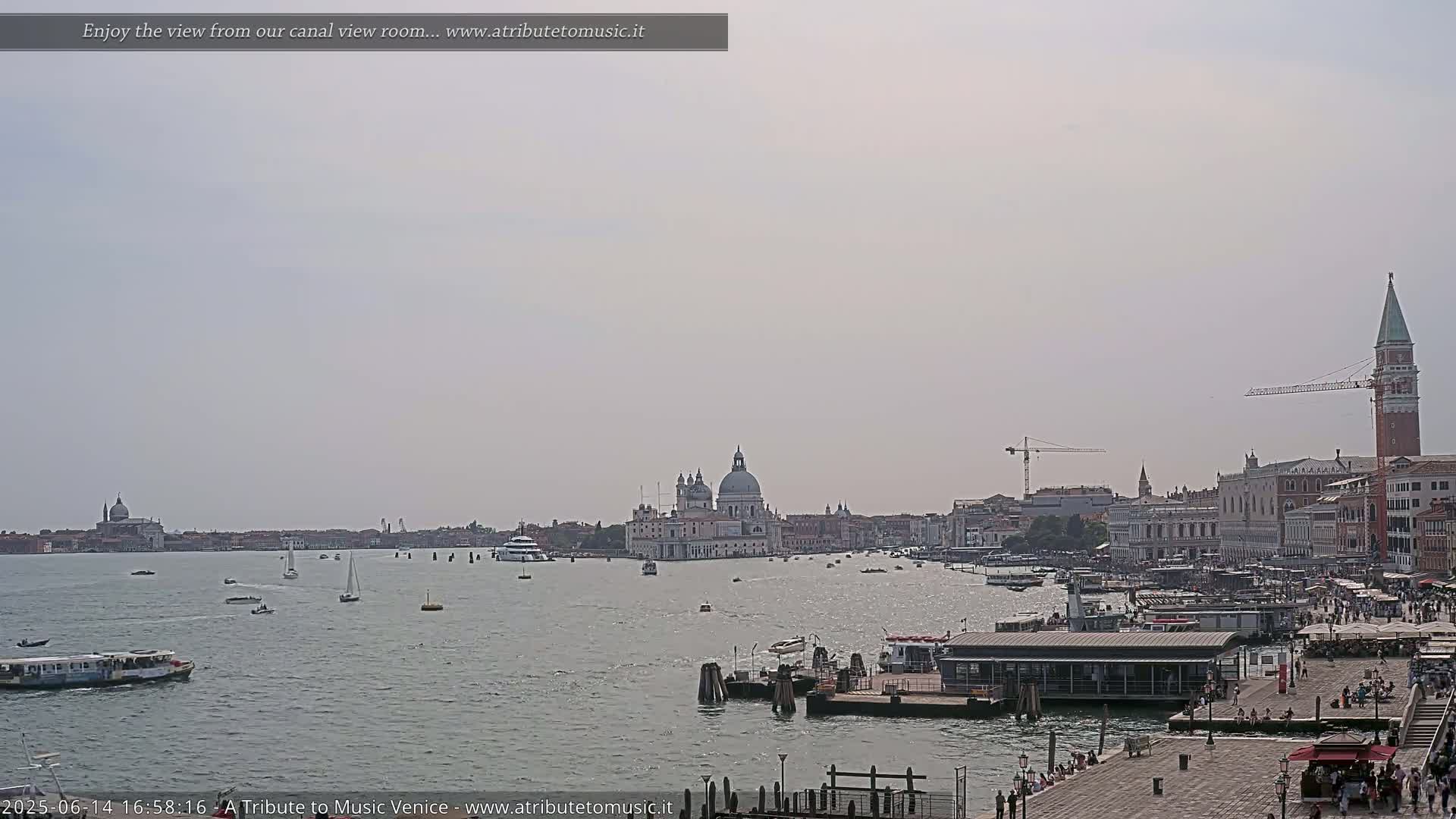 A hazy, sunny day reveals a wide Venetian canal with boats, a waterfront city featuring a large church and bell tower, and construction cranes in the distance.