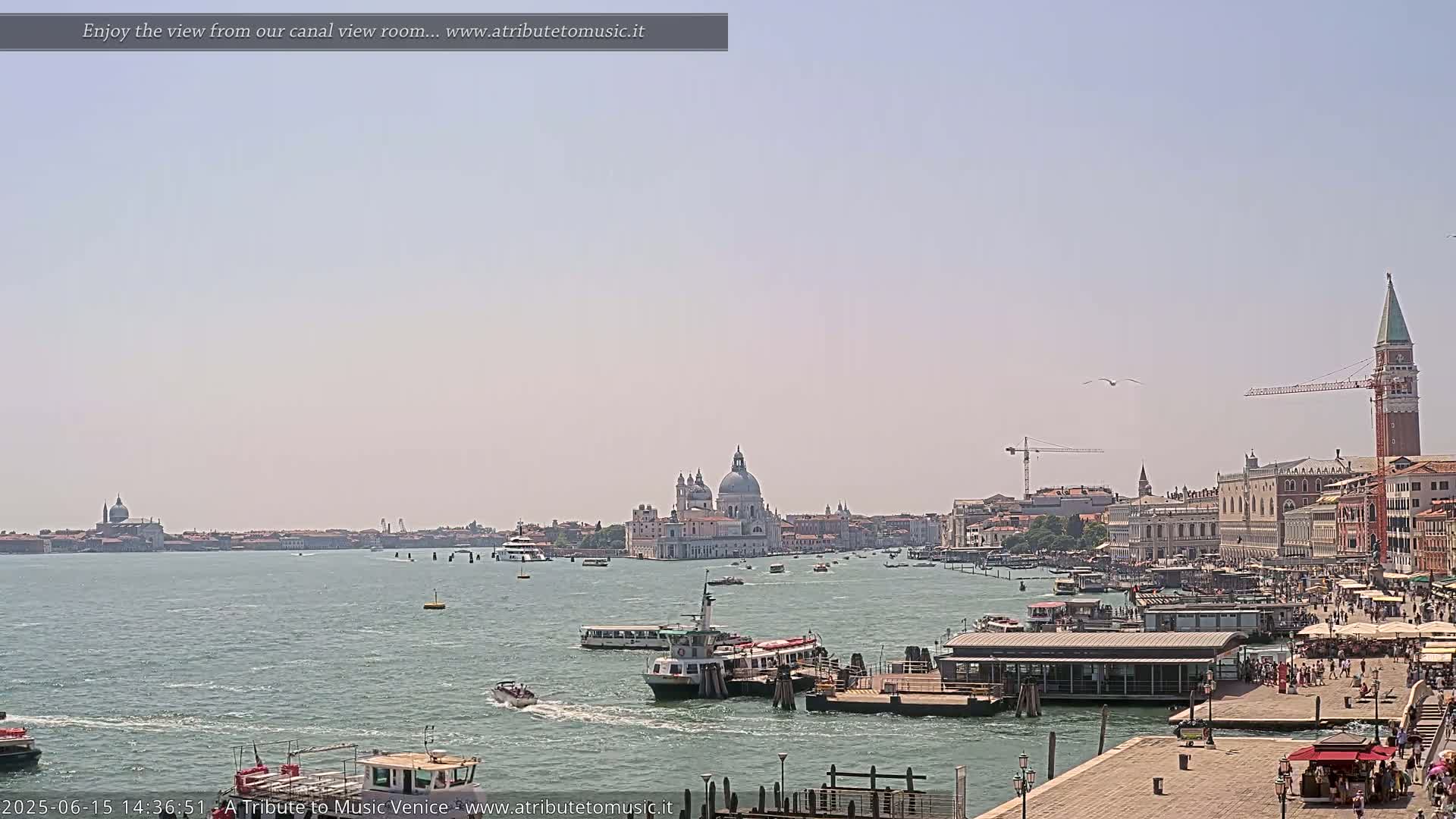 A sunny view across a calm canal shows boats and a Venetian cityscape with a large domed church and a tall bell tower.