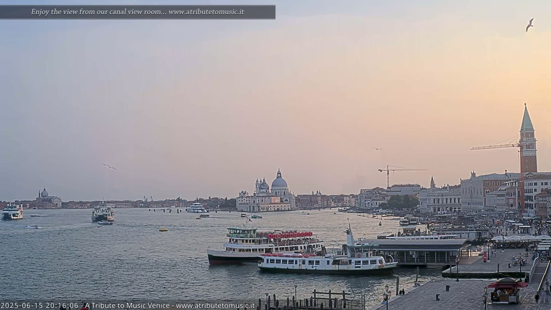 A calm canal in Venice, Italy at sunset, with several water taxis and boats, and a cityscape featuring a large church and the bell tower of St. Mark's Campanile in the background.