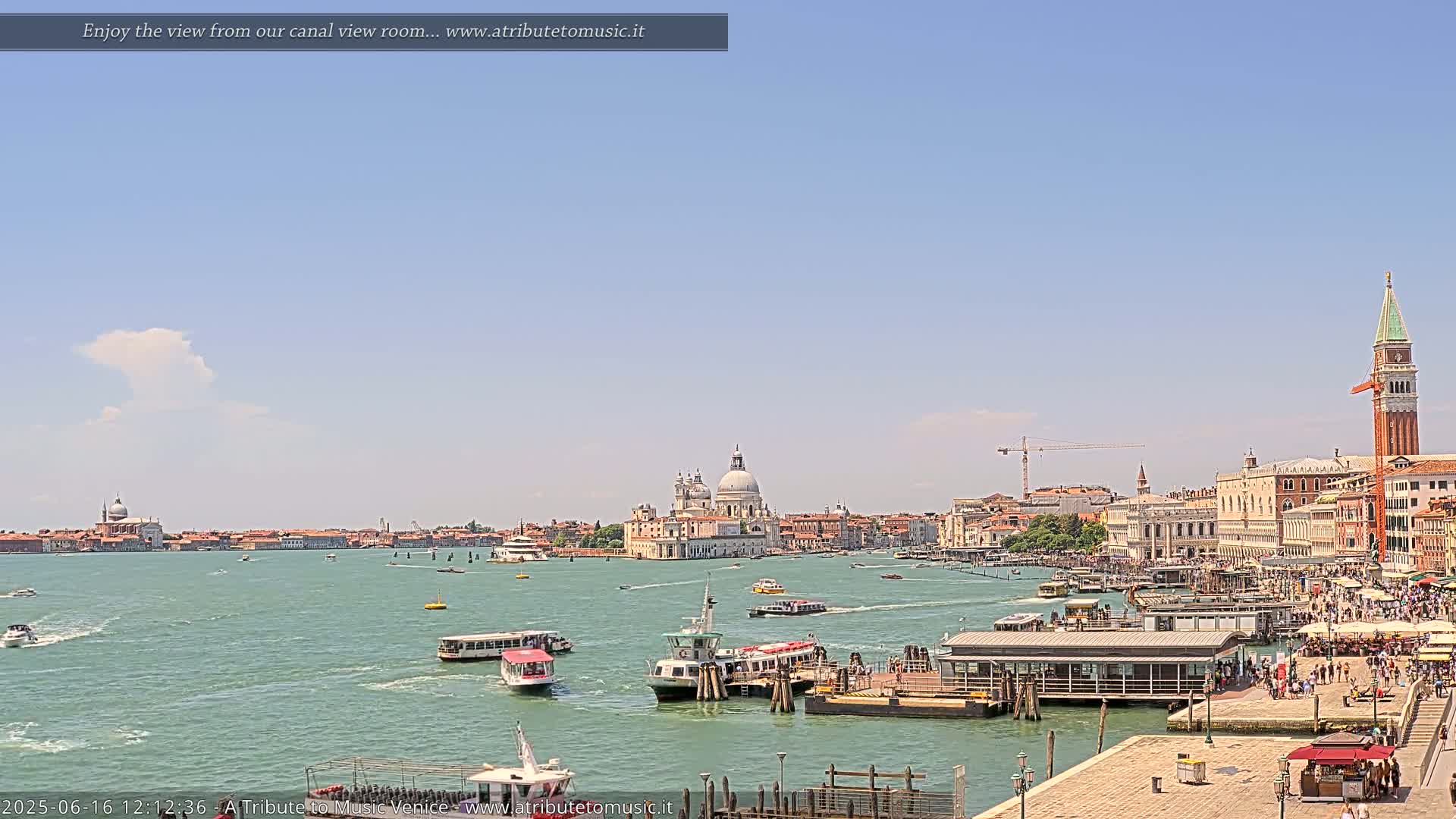 A sunny Venetian cityscape with numerous boats on a calm waterway, featuring a large basilica in the distance and a bell tower near the foreground.