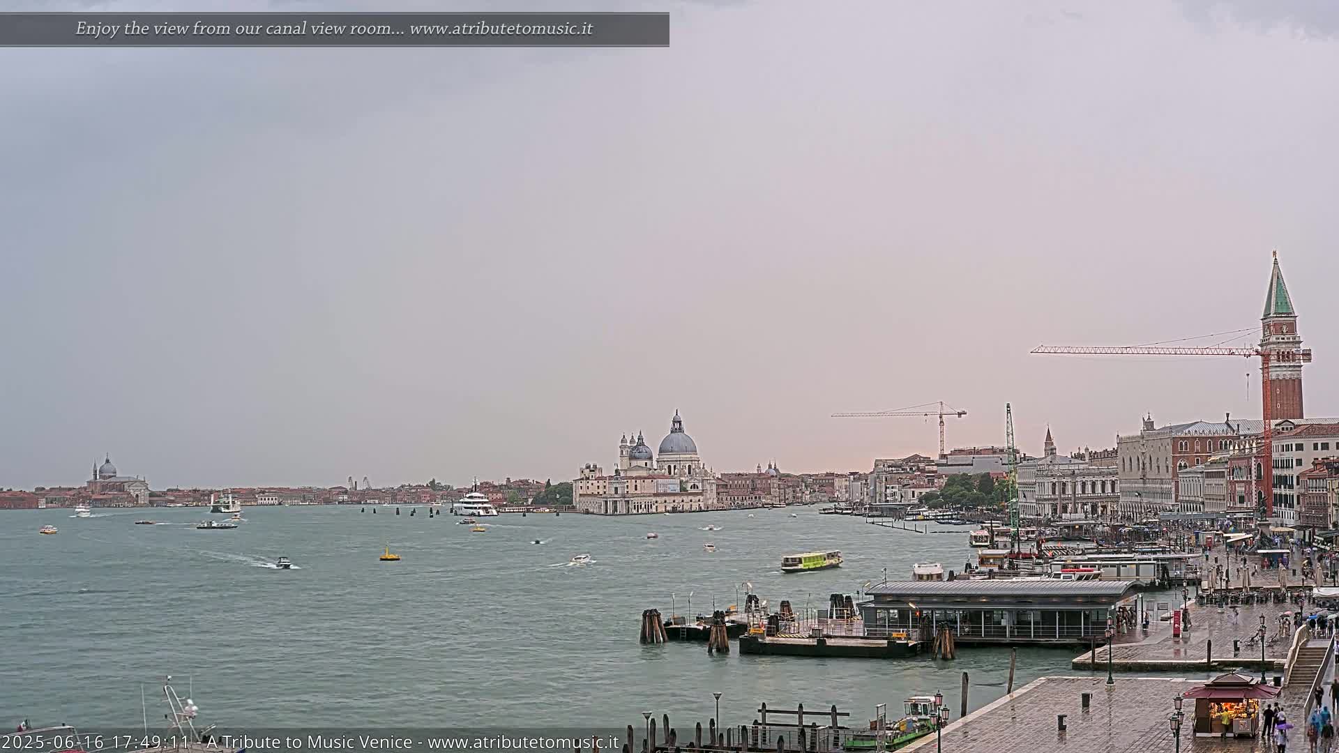 A wide shot of a Venetian canal on a cloudy day shows several boats, a waterfront building, and the Basilica di Santa Maria della Salute in the distance.
