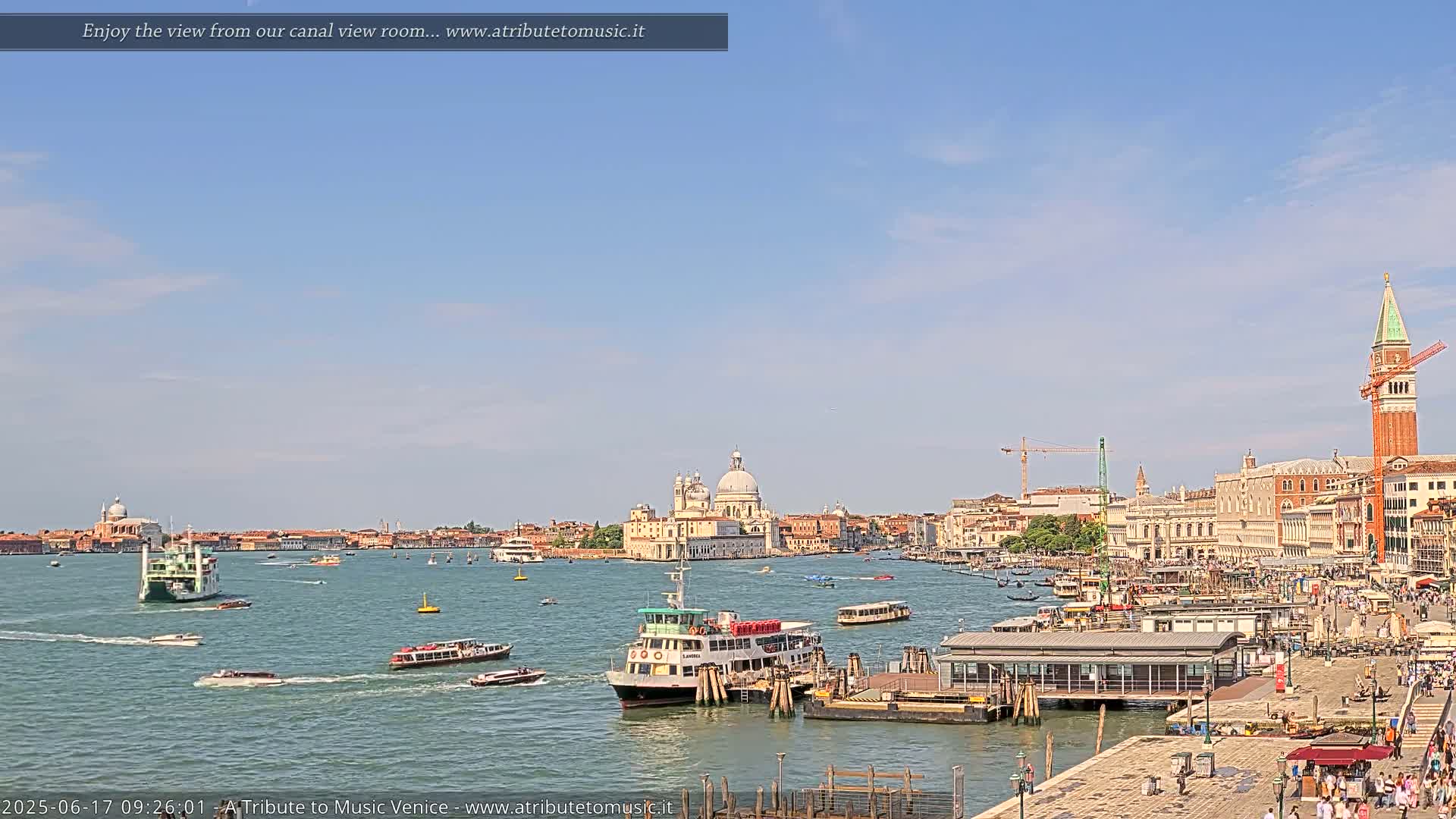 A sunny day in Venice shows a wide canal with numerous boats, including water taxis and ferries, and a cityscape featuring a large church and the bell tower of St. Mark's Campanile.