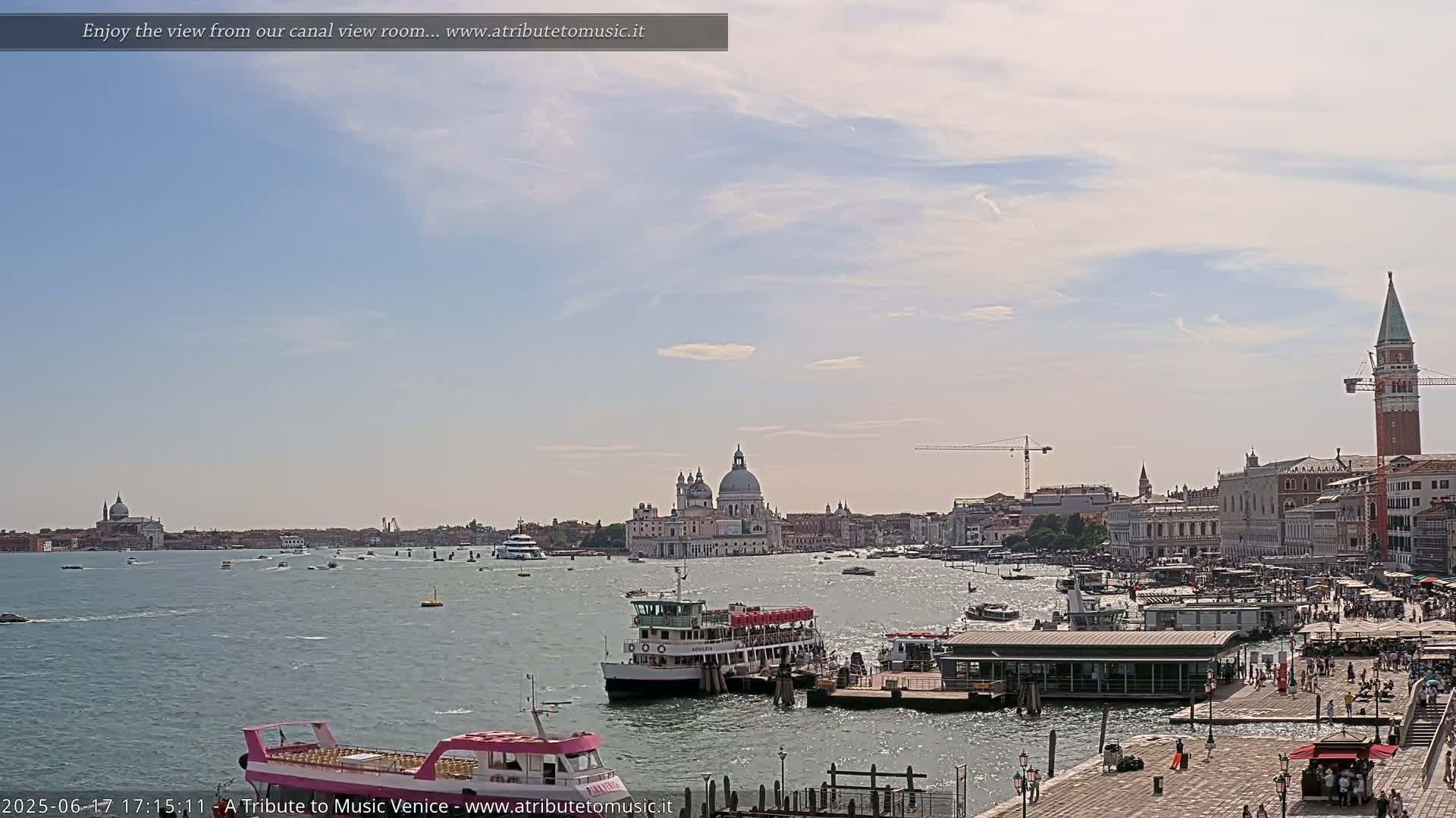 A wide shot of a Venetian canal on a partly cloudy day shows several boats, a waterfront promenade with people, and buildings in the distance, including a large domed church.