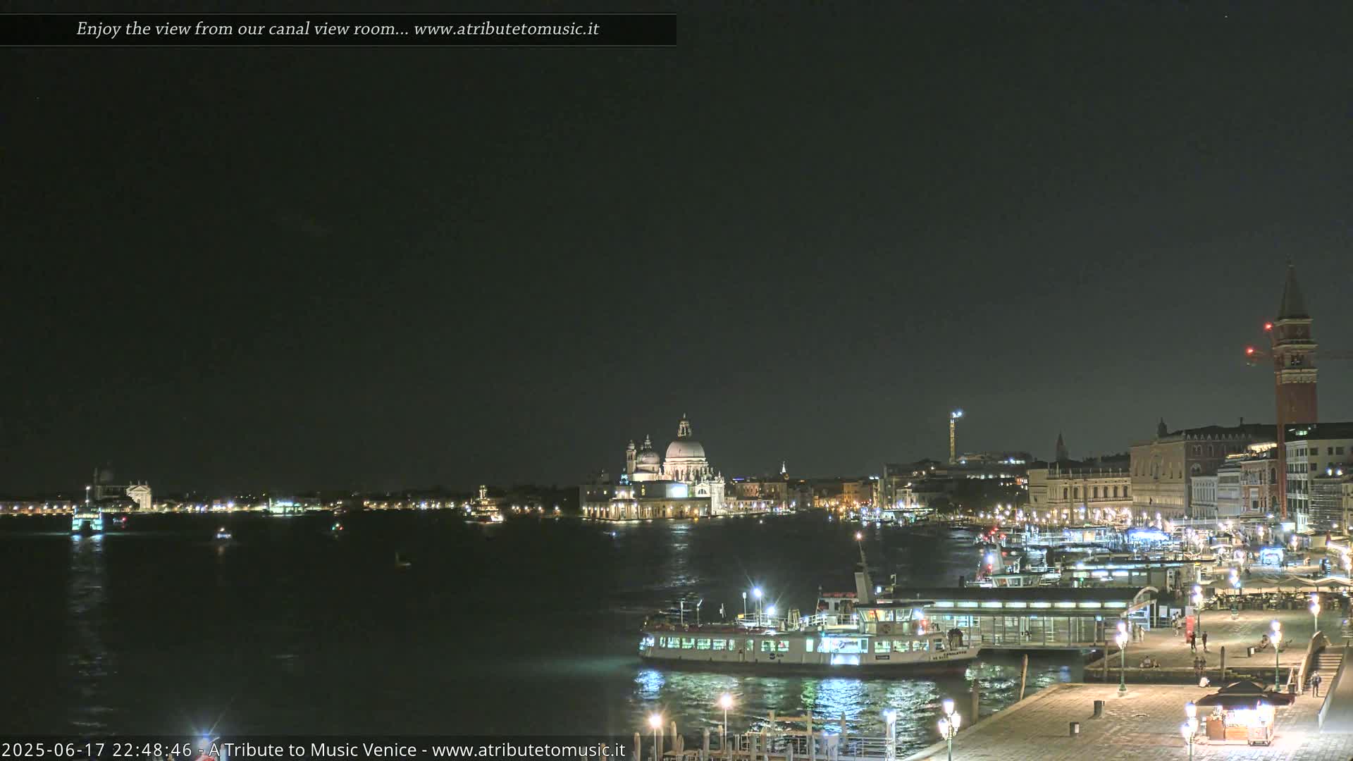 A nighttime view of a city waterfront with a large body of water, boats, and illuminated buildings, including a prominent domed structure.