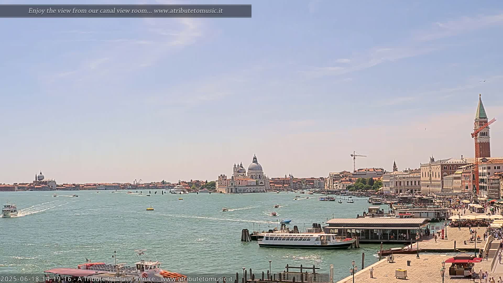 A wide shot of a waterway with numerous boats, showcasing a cityscape featuring a large church and tall bell tower under a mostly clear sky.
