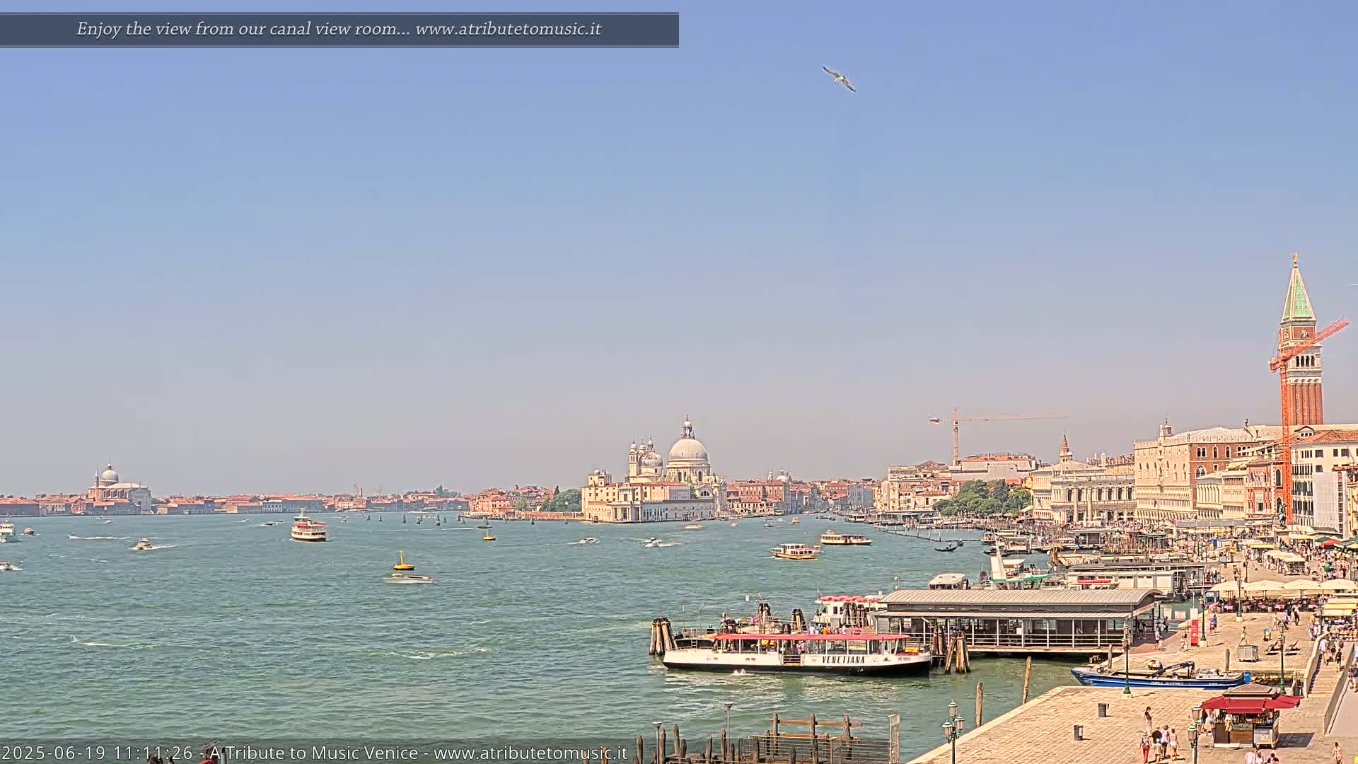 A sunny Venetian cityscape with a large body of water showing various boats, including a ferry at a dock, with the Basilica di Santa Maria della Salute prominently featured in the distance.
