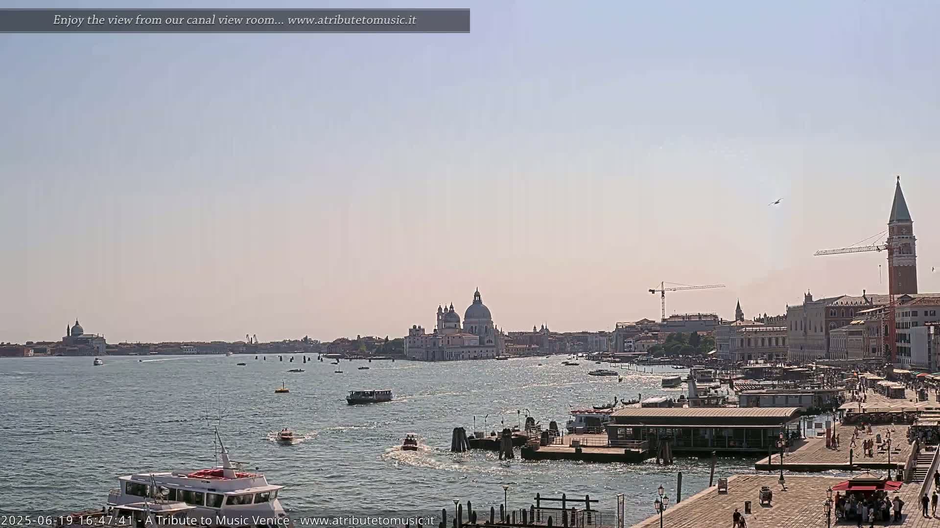A sunny Venetian canal scene shows boats on the water, with buildings and a large church in the distance.