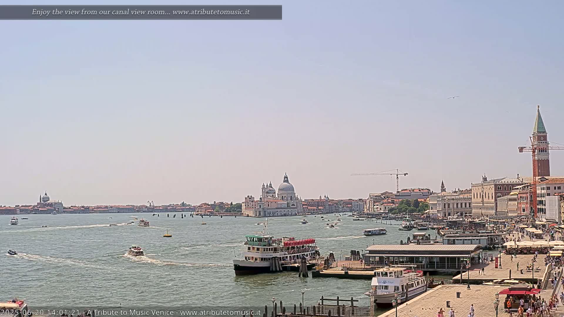 A sunny day in Venice shows a waterway with several boats, including water taxis, and a view of the city skyline including the Basilica di Santa Maria della Salute and St. Mark's Campanile.
