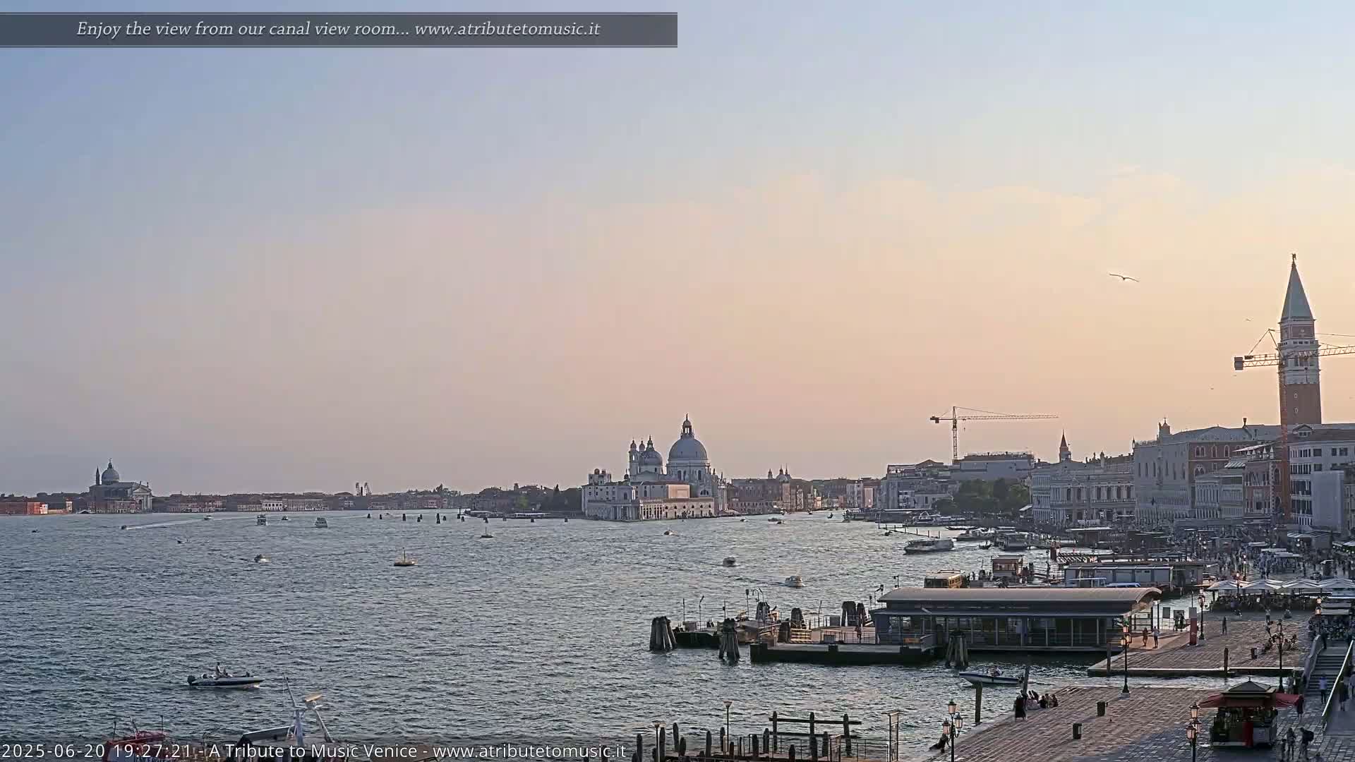 A wide shot of a calm Venetian waterway at sunset, showing boats, buildings, and a distant church dome under a pale sky.