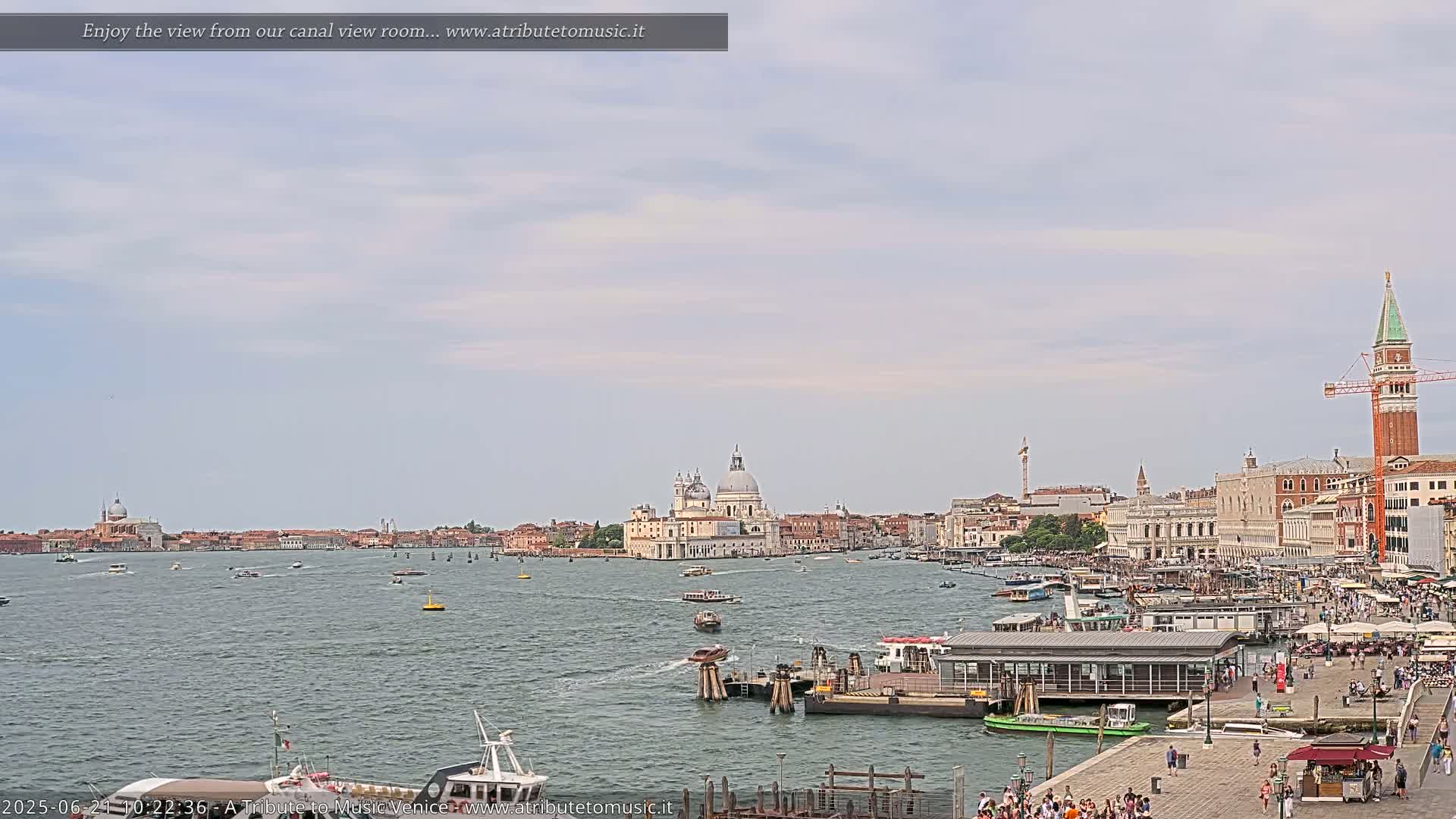 A wide shot of a canal in Venice, Italy on a partly cloudy day, shows numerous boats and buildings lining the waterway, including a large church with a dome.