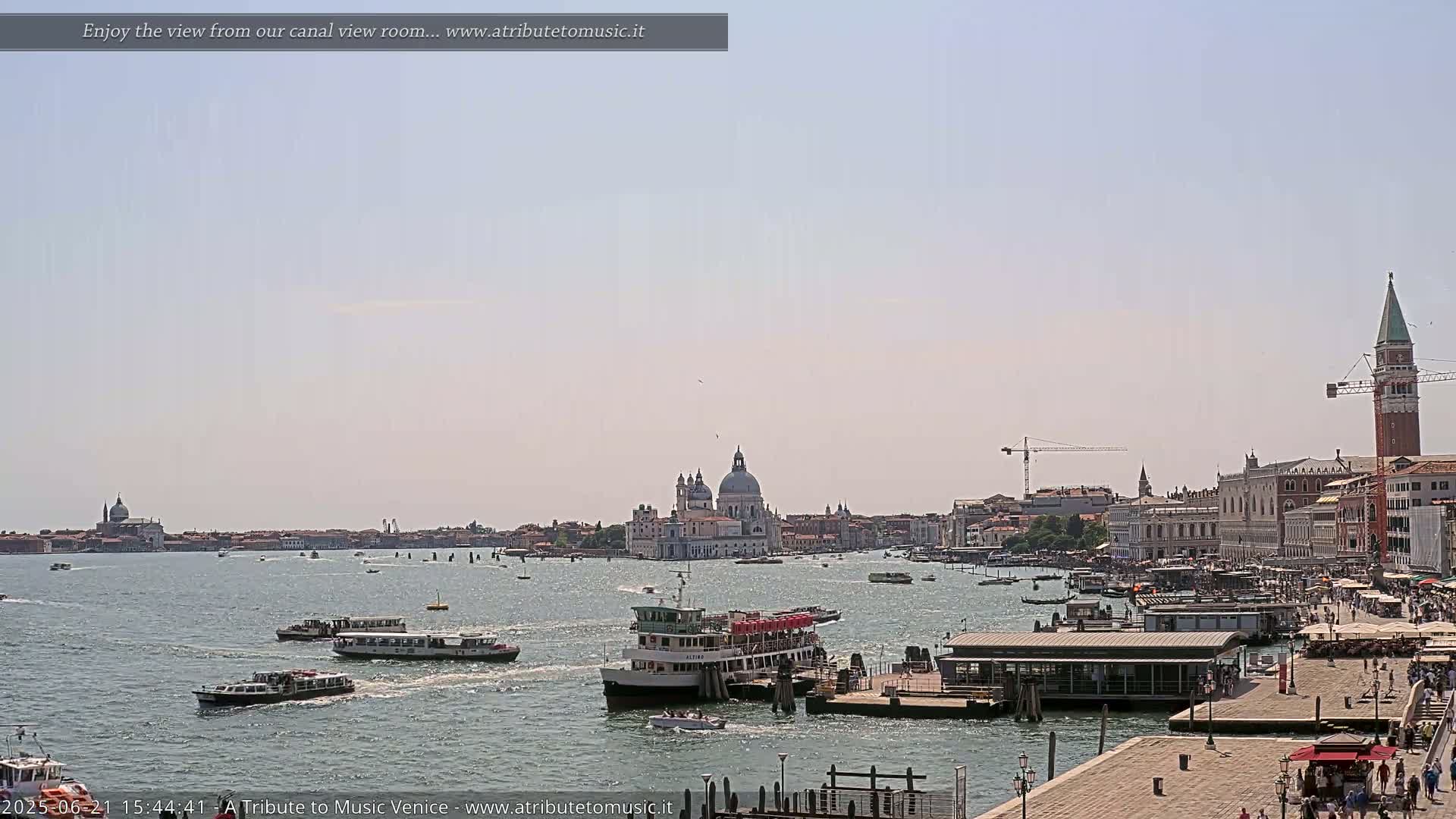A sunny day in Venice shows several water taxis and ferries navigating a canal toward a cityscape featuring a large domed church and a tall bell tower.