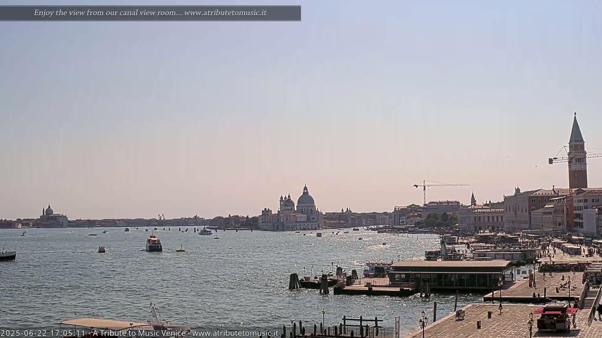 A wide view of a Venetian canal shows boats, buildings, and a distant church under a clear, bright sky.