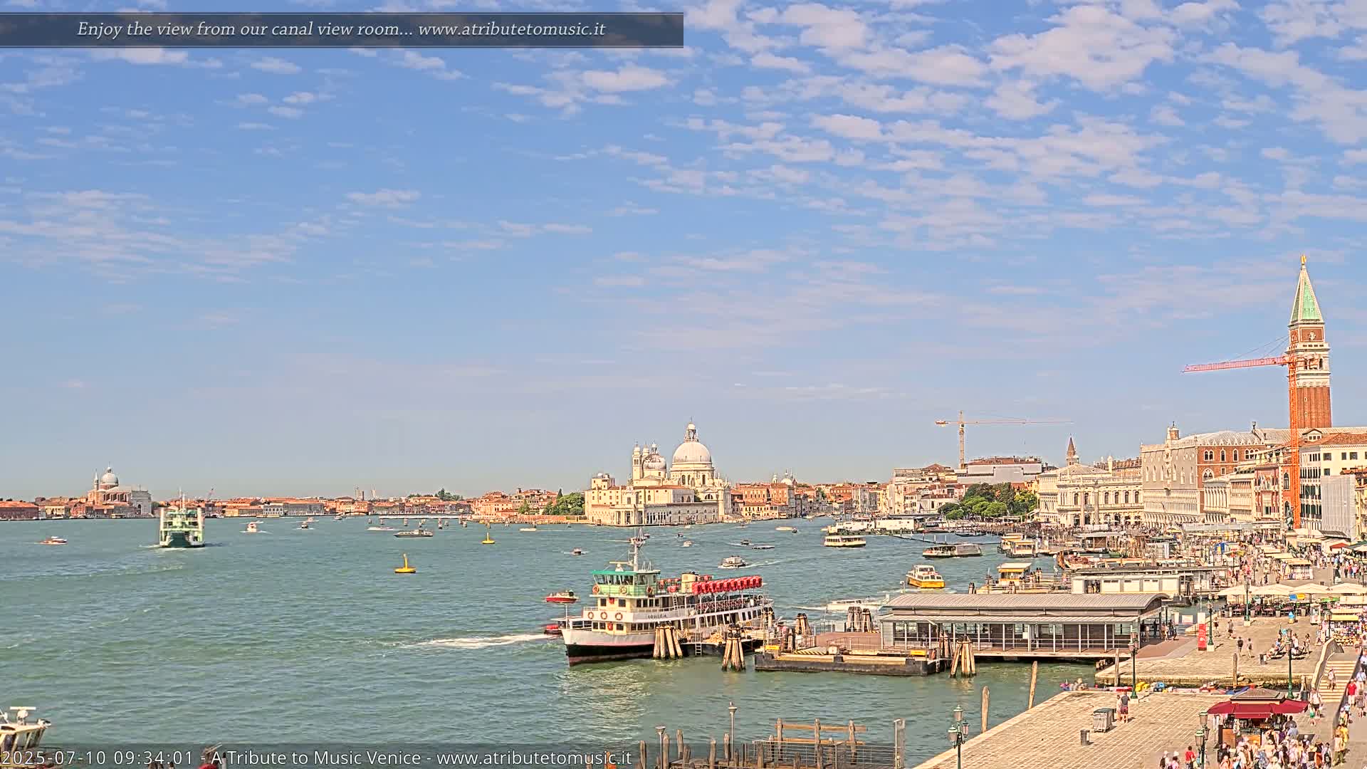 A sunny day in Venice shows a wide canal with several boats, a ferry docked at a pier, and a cityscape including a large domed church and the bell tower of St. Mark's Square in the distance.