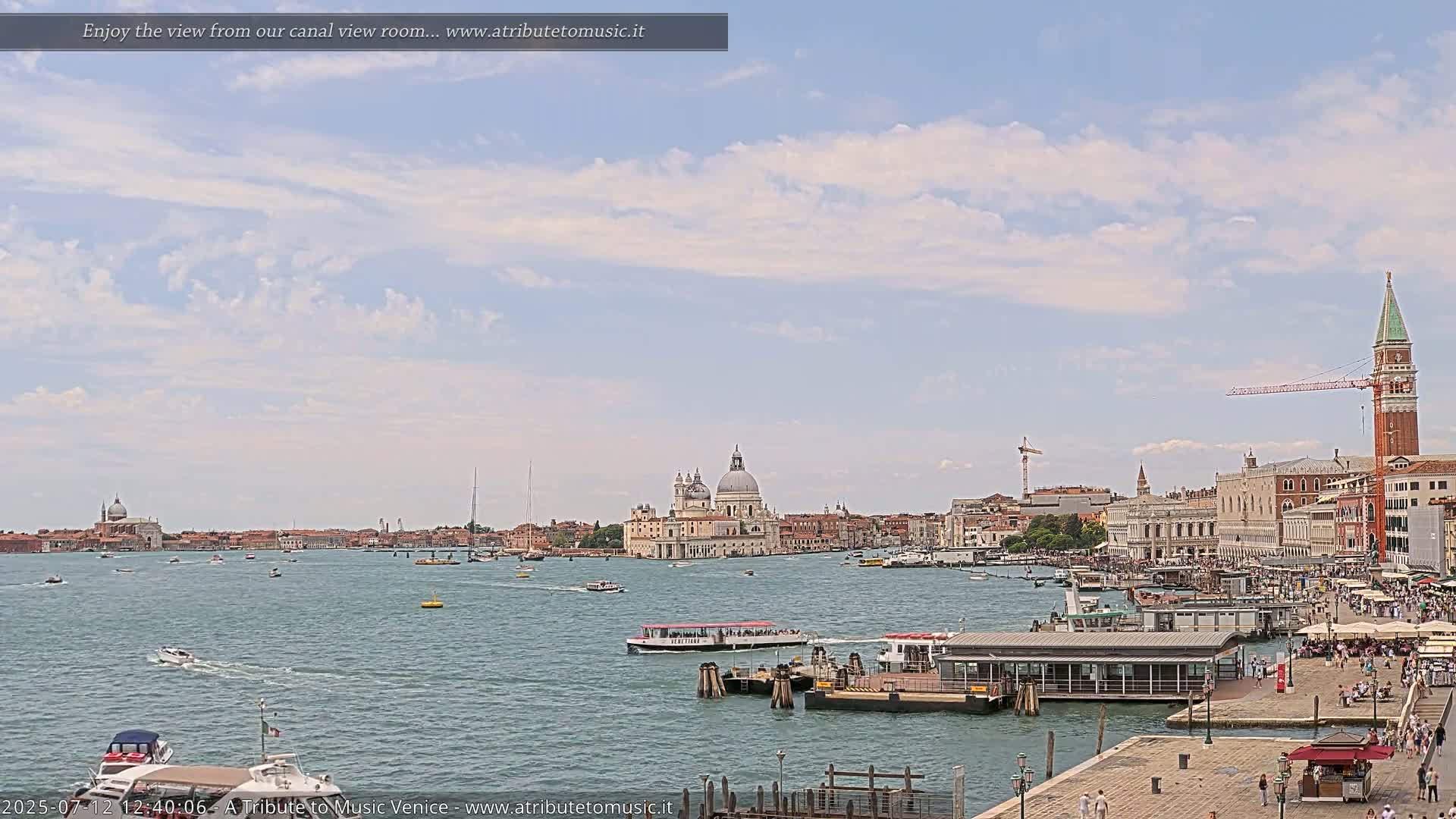 A sunny day in Venice shows a wide canal with several boats, a ferry docked at a pier, and a cityscape including a large domed church and the bell tower of St. Mark's Square in the distance.