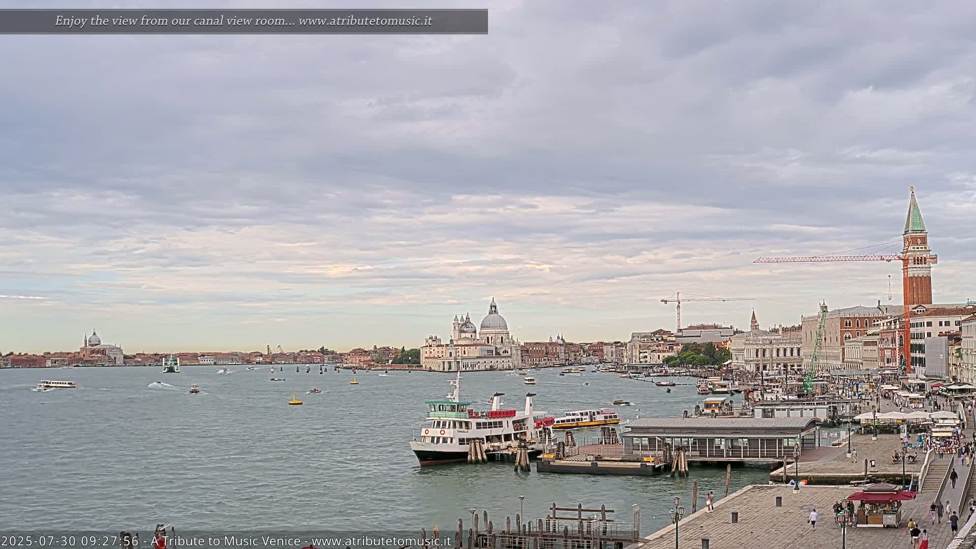 A wide view of a Venetian canal shows several boats, a ferry at a dock, and buildings along the waterfront under a mostly cloudy sky.