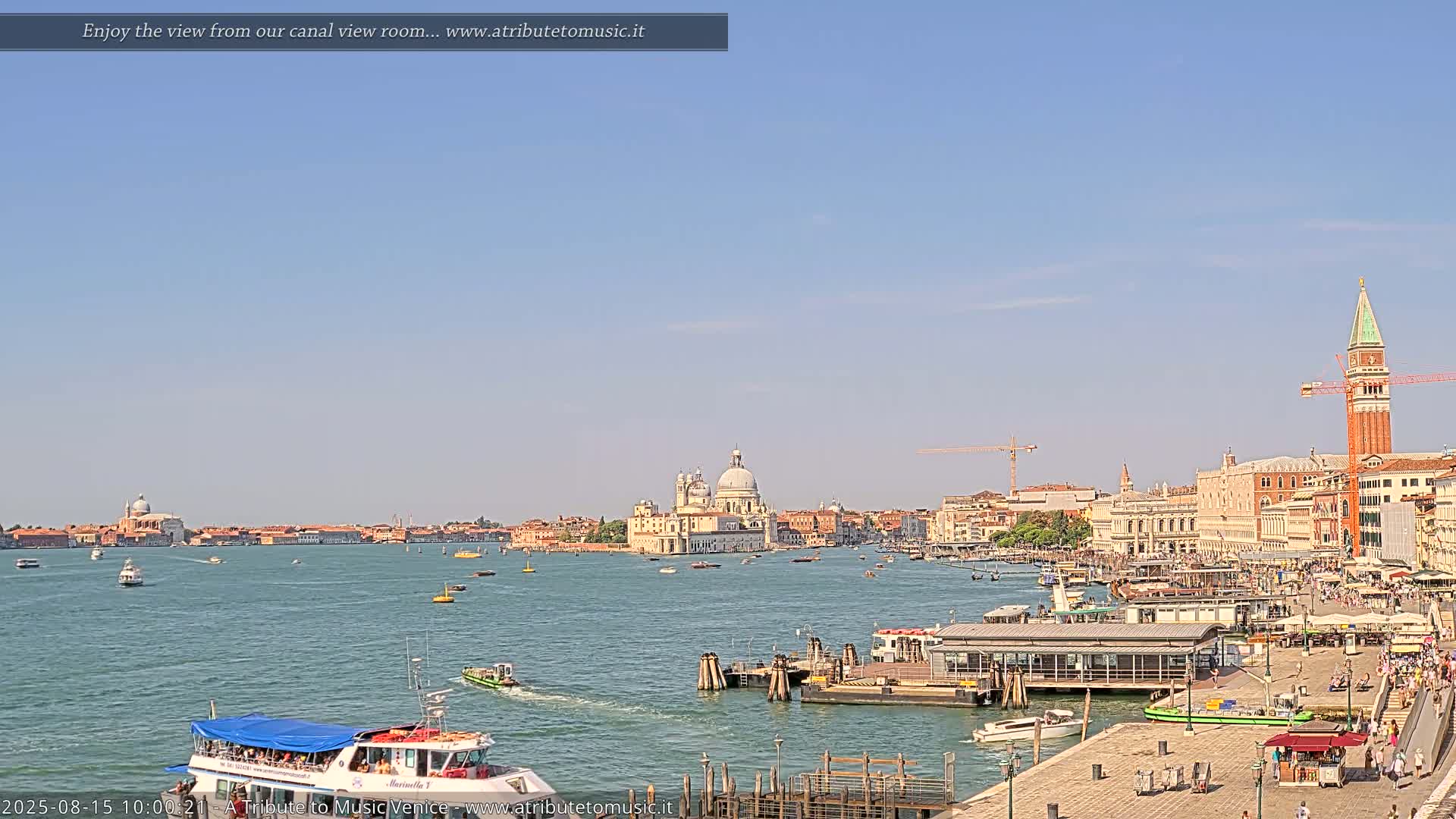 A sunny day in Venice shows a wide canal with boats, a waterfront with people, and buildings including a large church and a tall tower in the background.