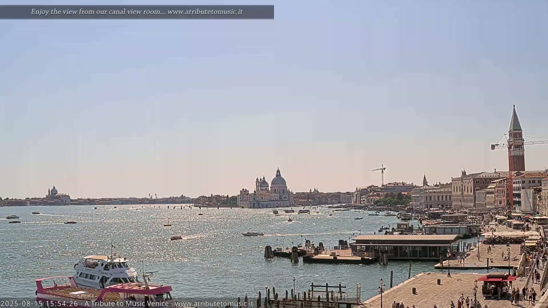 A sunny day in Venice shows a wide canal with boats, a waterfront with people, and buildings including a large church and a tall tower in the background.