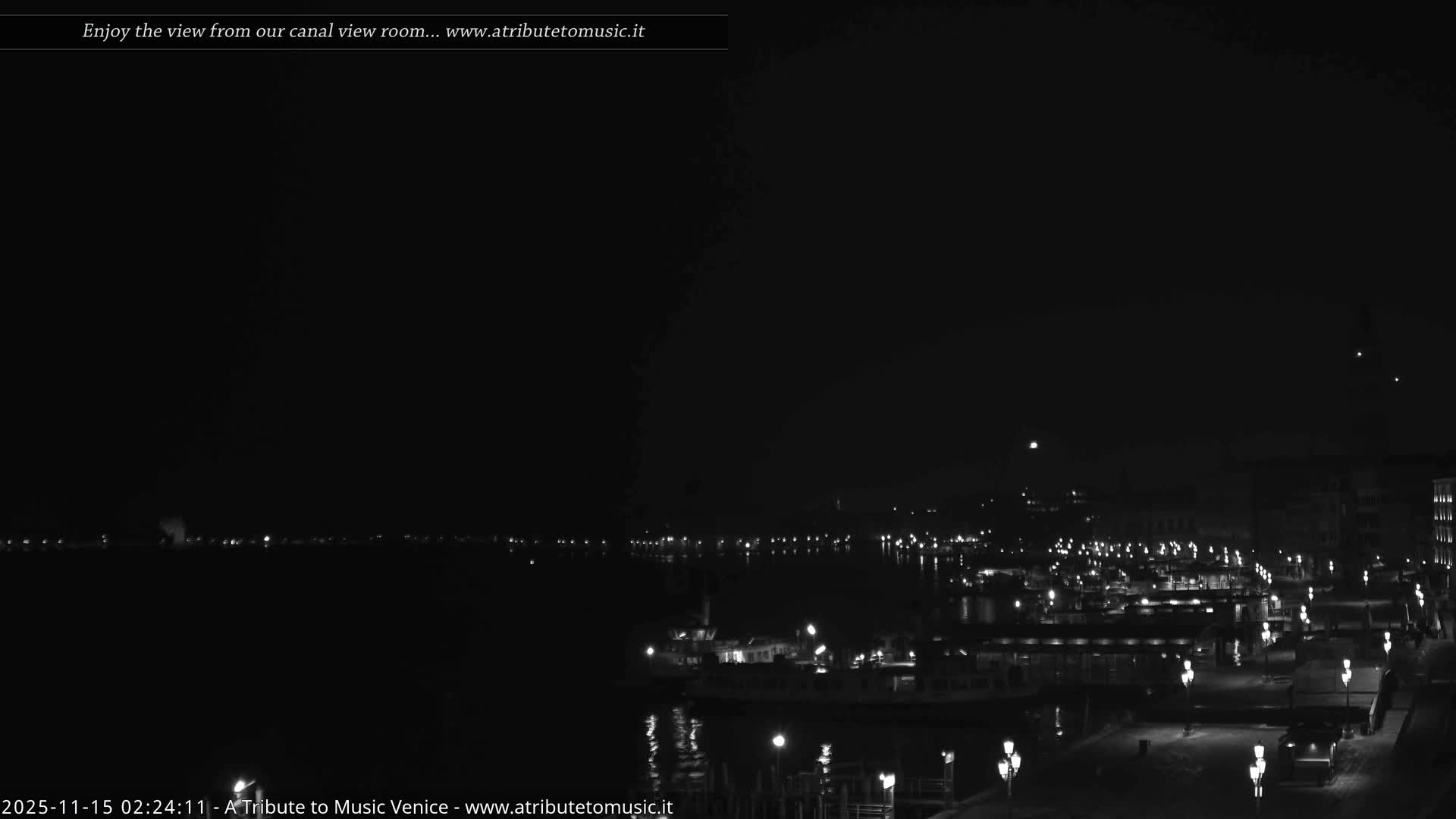 The image displays a clear night scene of a calm body of water with boats docked along a waterfront, illuminated by numerous city lights reflecting on the surface, under a dark, clear sky.