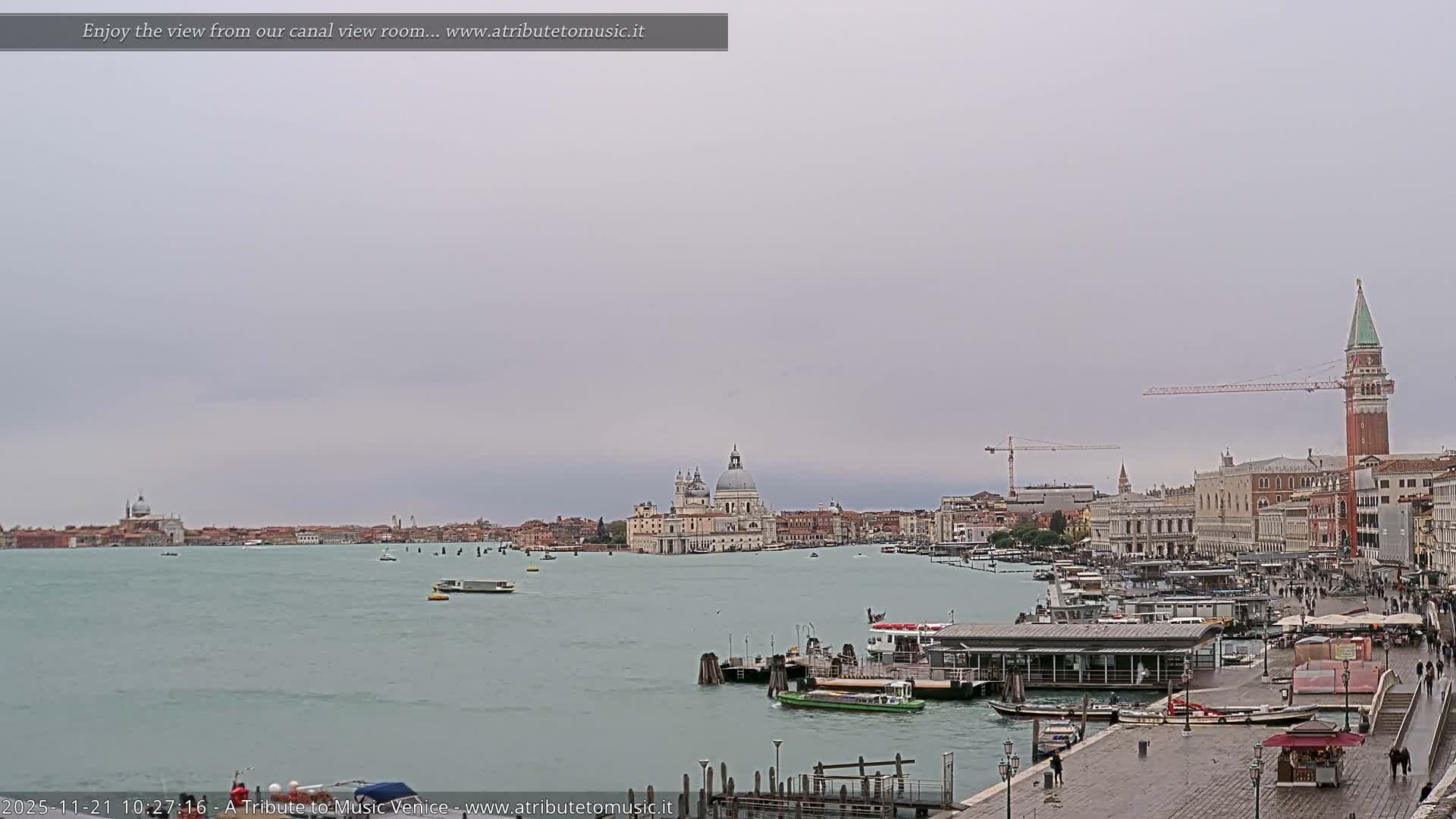 On an overcast day, a wide view of Venice captures numerous boats on a calm canal flanked by historic buildings, including Santa Maria della Salute and St Mark's Campanile, alongside a bustling waterfront promenade.