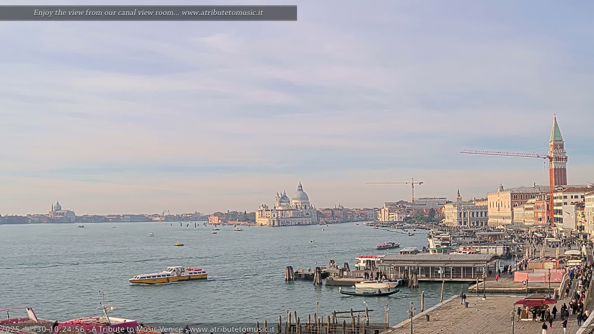 A wide panoramic view of Venice's Grand Canal on a partly cloudy and bright day, features numerous boats navigating the water, the prominent domes of Santa Maria della Salute, the towering St. Mark's Campanile, and a bustling waterfront promenade lined with historic buildings and people.