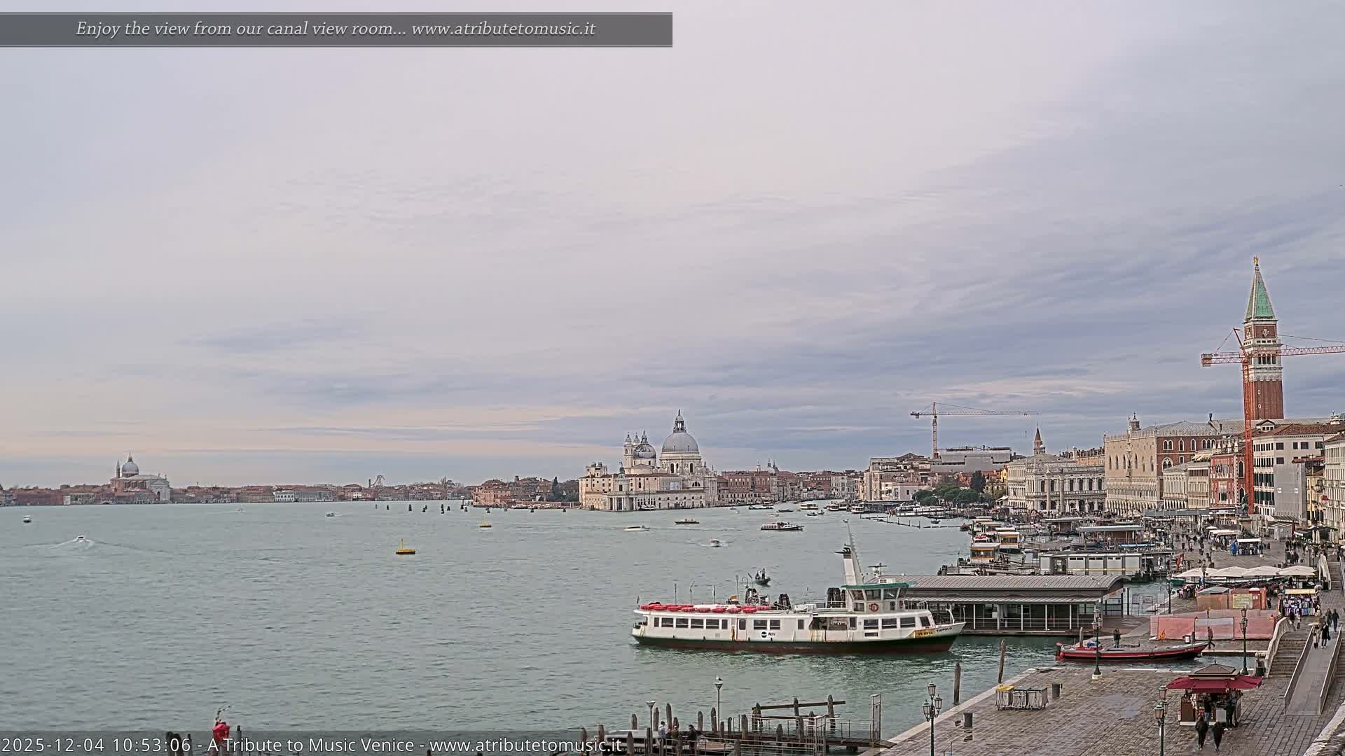 A wide panoramic view of Venice on an overcast day shows the busy waterway filled with boats, a lively waterfront promenade, and the city's iconic architecture, including the Santa Maria della Salute basilica and St. Mark's Campanile, under a cloudy sky.