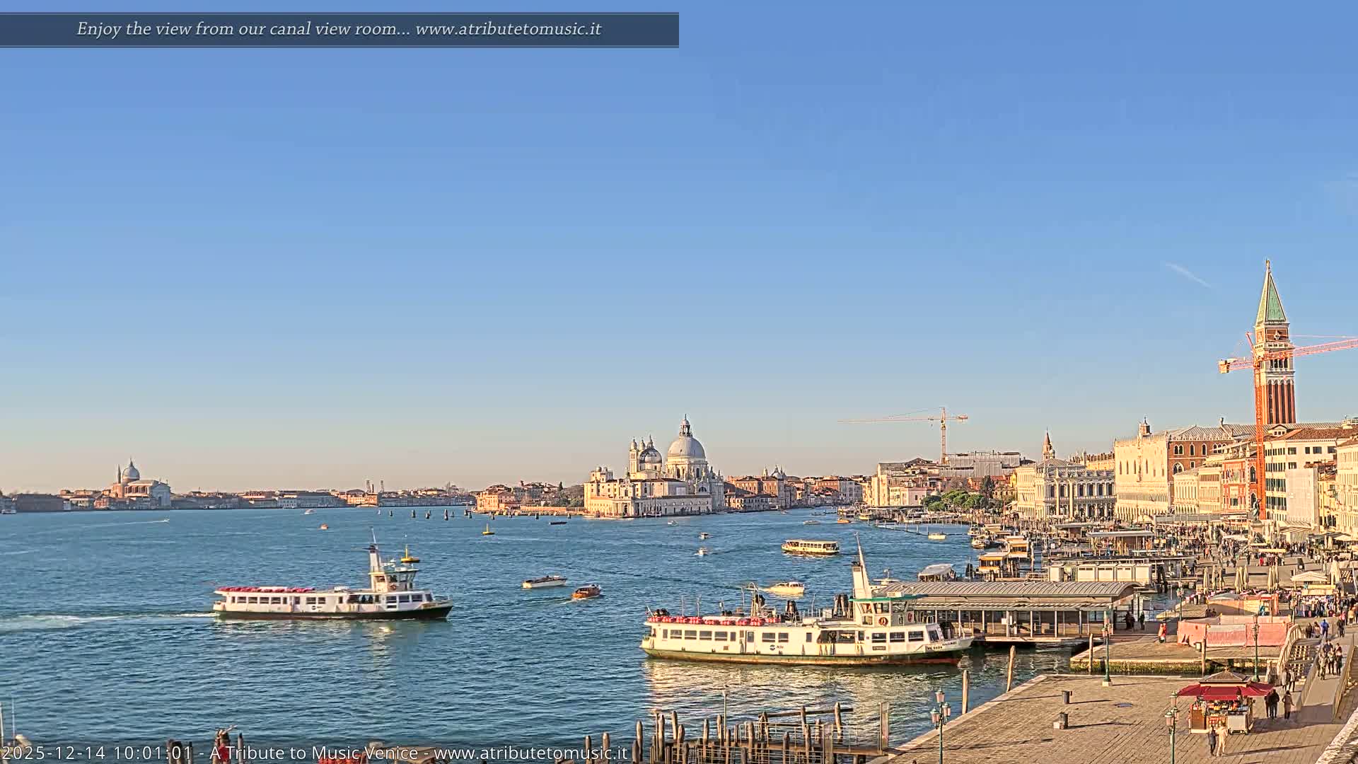 A wide panoramic view of Venice on an overcast day shows the busy waterway filled with boats, a lively waterfront promenade, and the city's iconic architecture, including the Santa Maria della Salute basilica and St. Mark's Campanile, under a cloudy sky.
