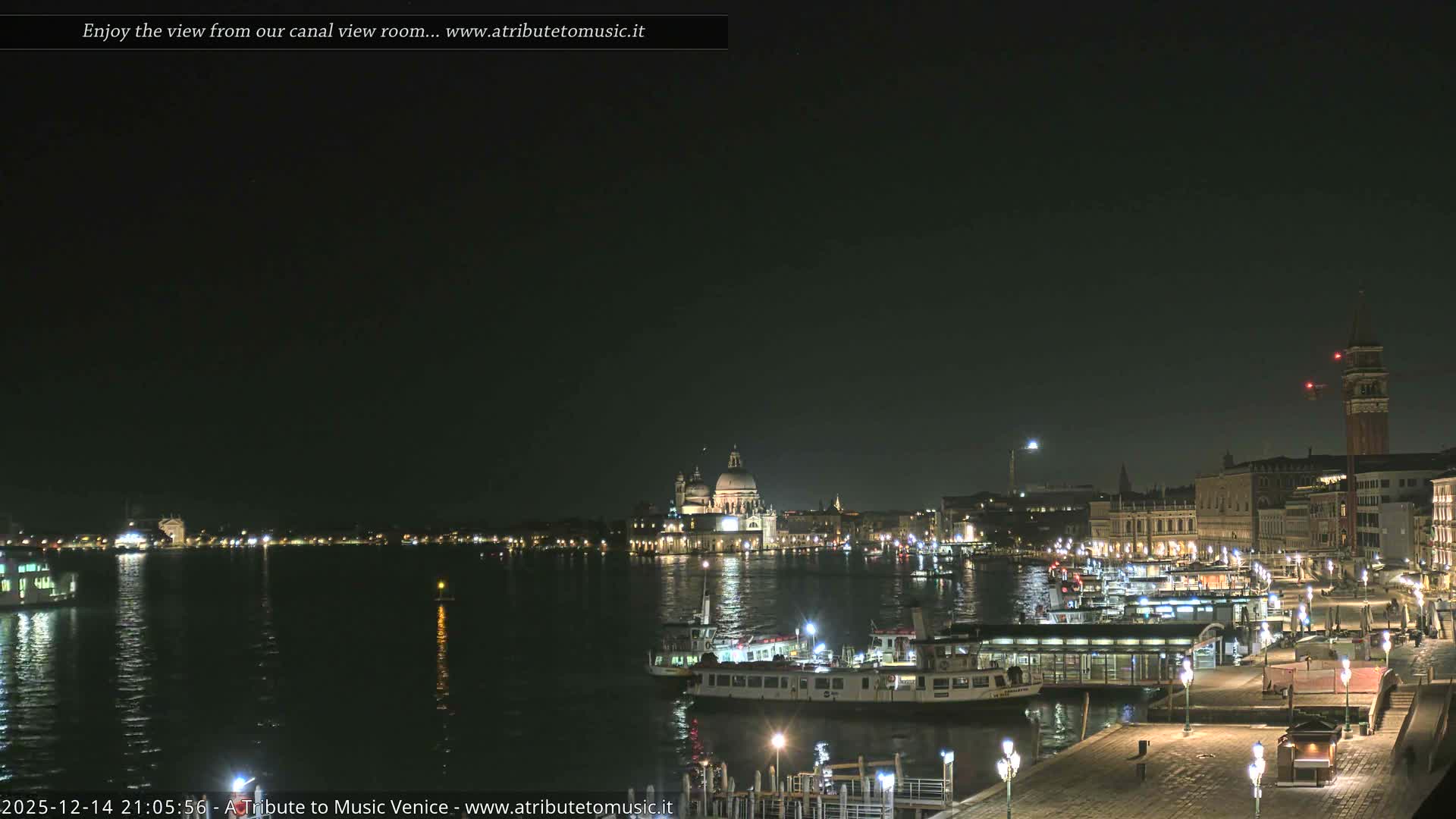 A wide panoramic view of Venice on an overcast day shows the busy waterway filled with boats, a lively waterfront promenade, and the city's iconic architecture, including the Santa Maria della Salute basilica and St. Mark's Campanile, under a cloudy sky.