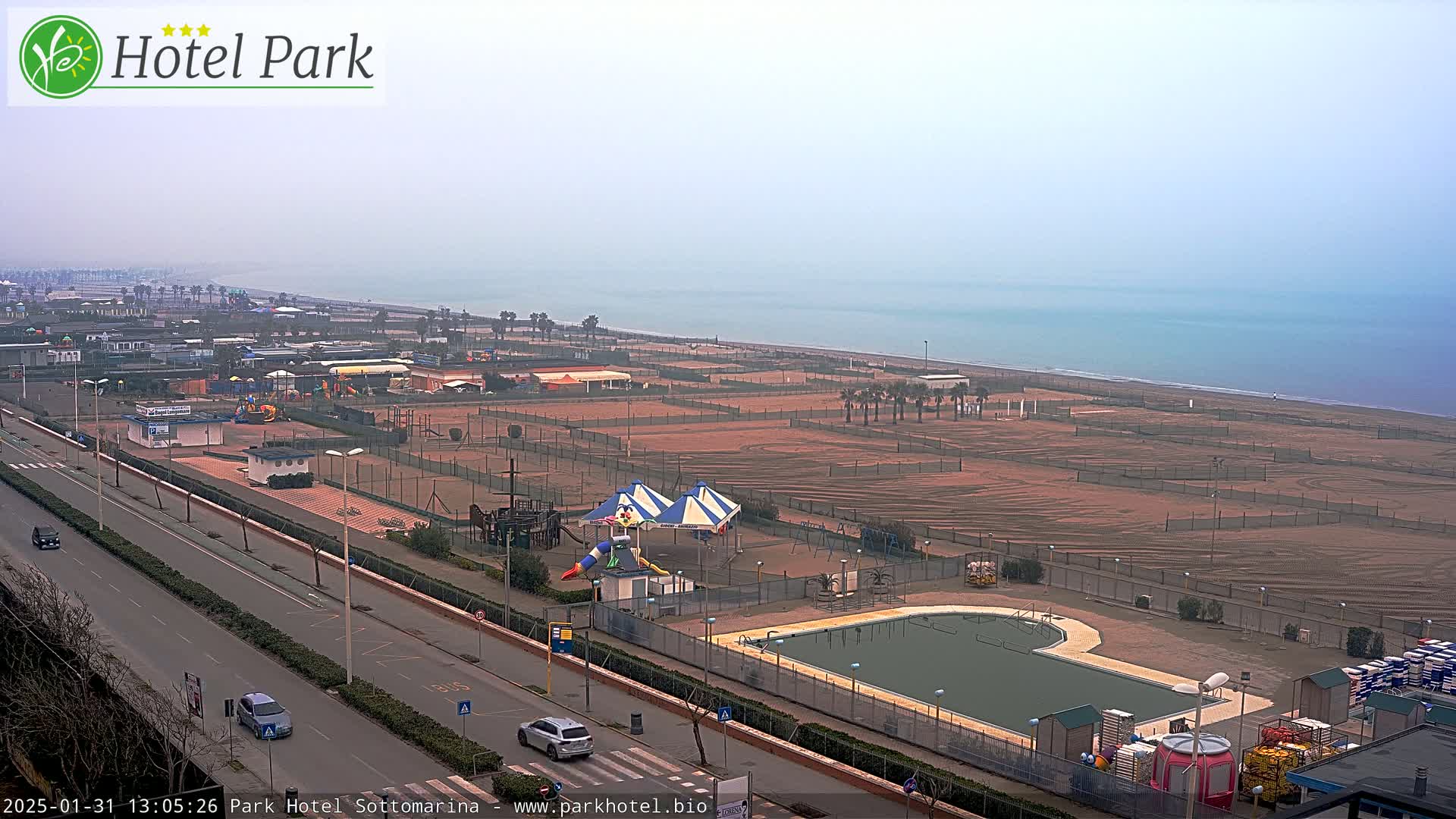 A hazy coastal view shows a sandy beach with volleyball courts, a playground, a swimming pool, and a road with cars.