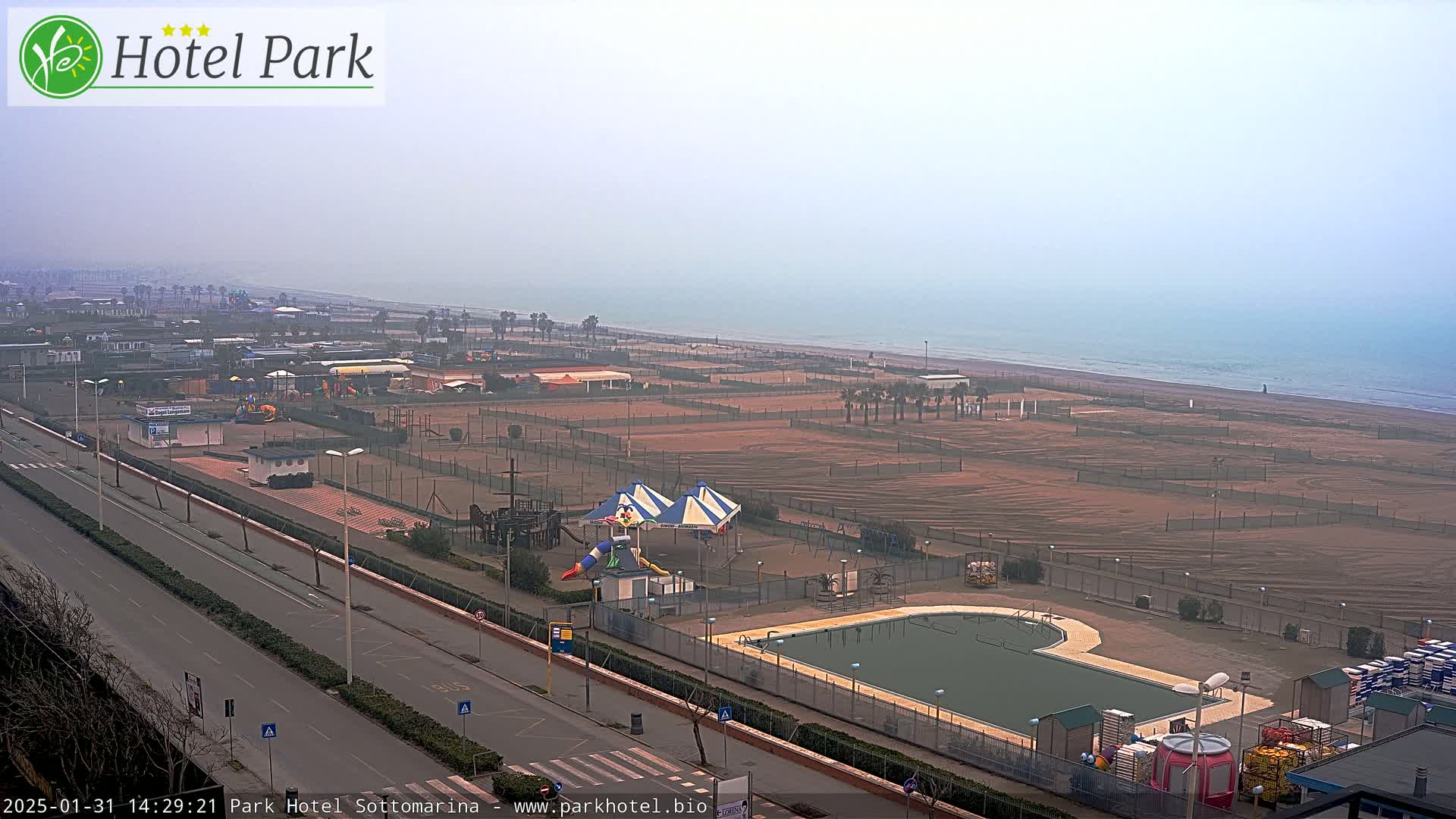 A hazy, overcast day reveals a beachside resort area with a swimming pool, playground, and empty sandy beach stretching to a calm sea.