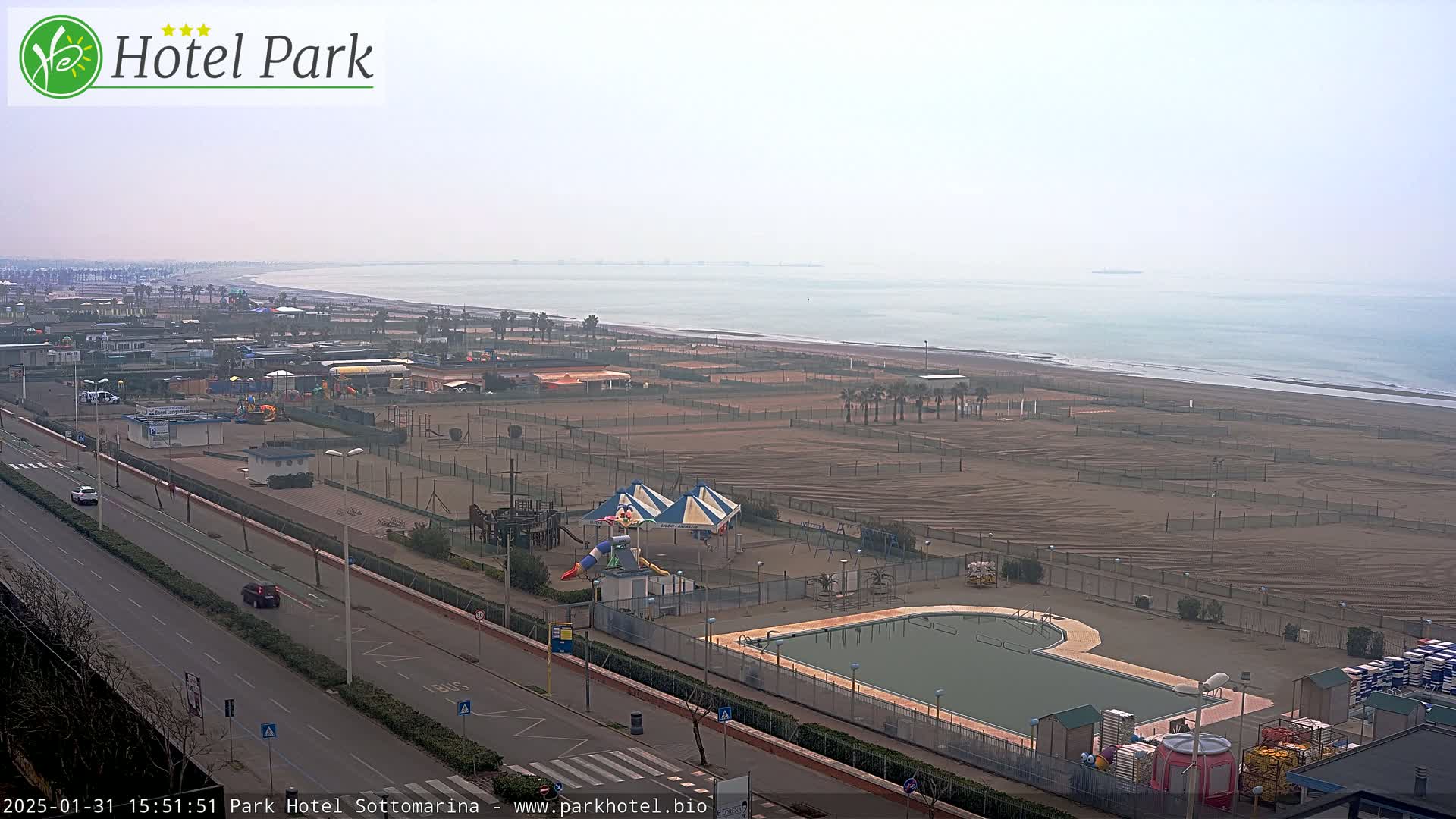 A hazy, overcast day reveals a coastal scene with a sandy beach, a swimming pool, playground equipment, and a road with sparse traffic.