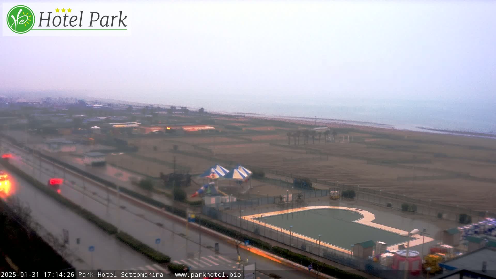 An overcast view shows a beach, a road with cars, a swimming pool, and playground equipment near a hotel.