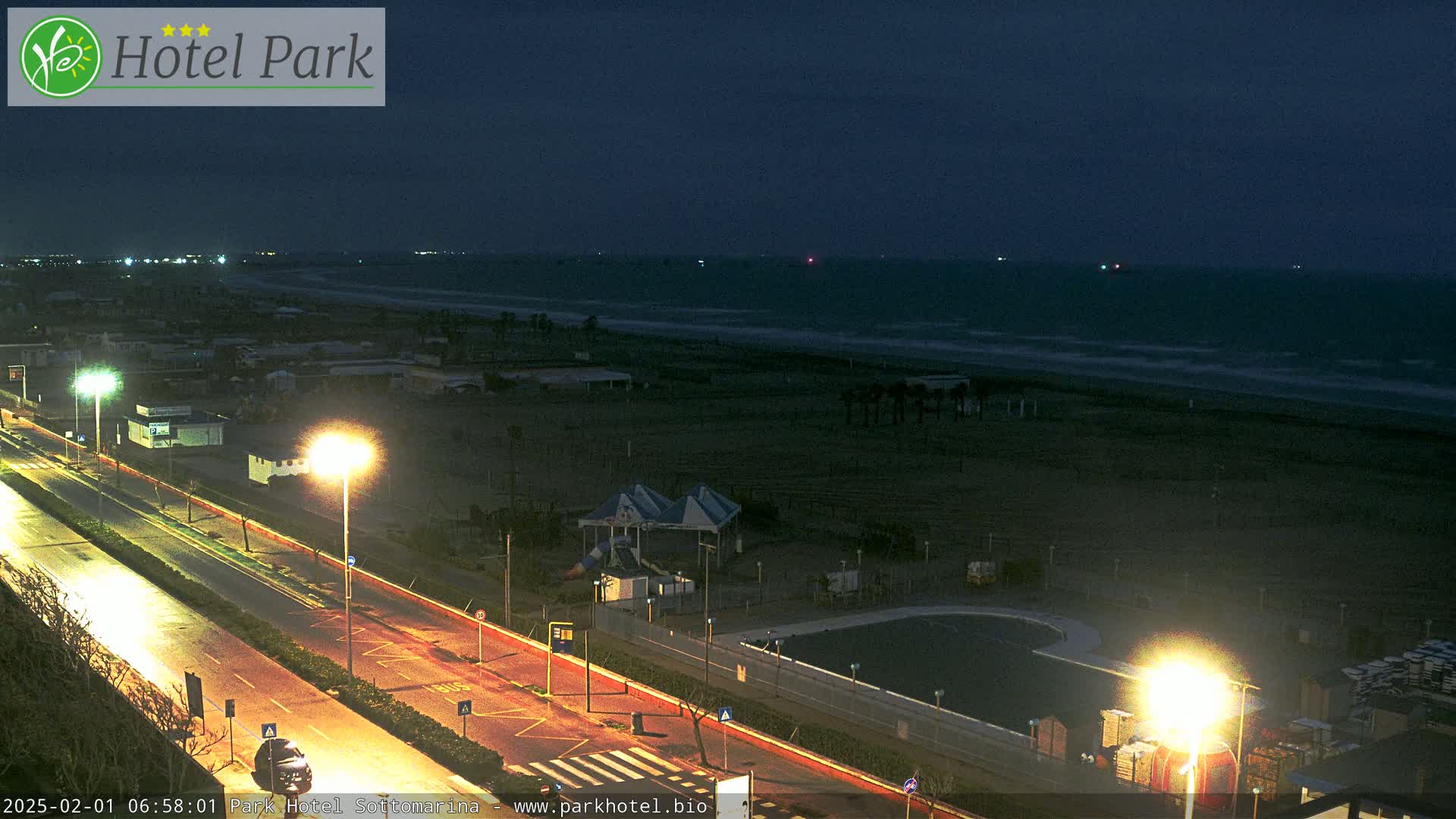 A nighttime view of a beach with a road, streetlights, and a small pool, under a dark, overcast sky.