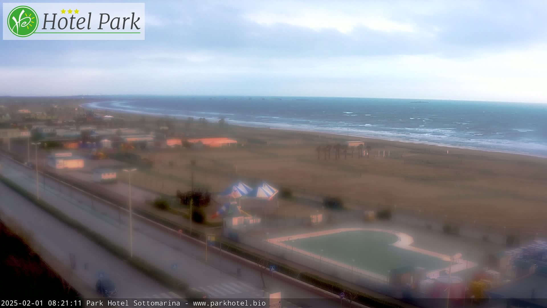 A beach with a wavy ocean under an overcast sky is viewed from above, showing a road and recreational areas nearby.
