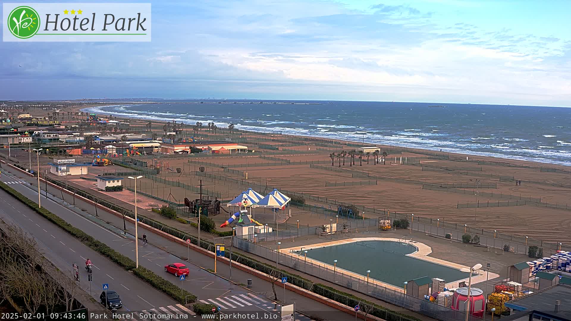 A seaside resort area with a playground, swimming pool, and sandy beach shows a partly cloudy sky and a windy ocean with small waves.