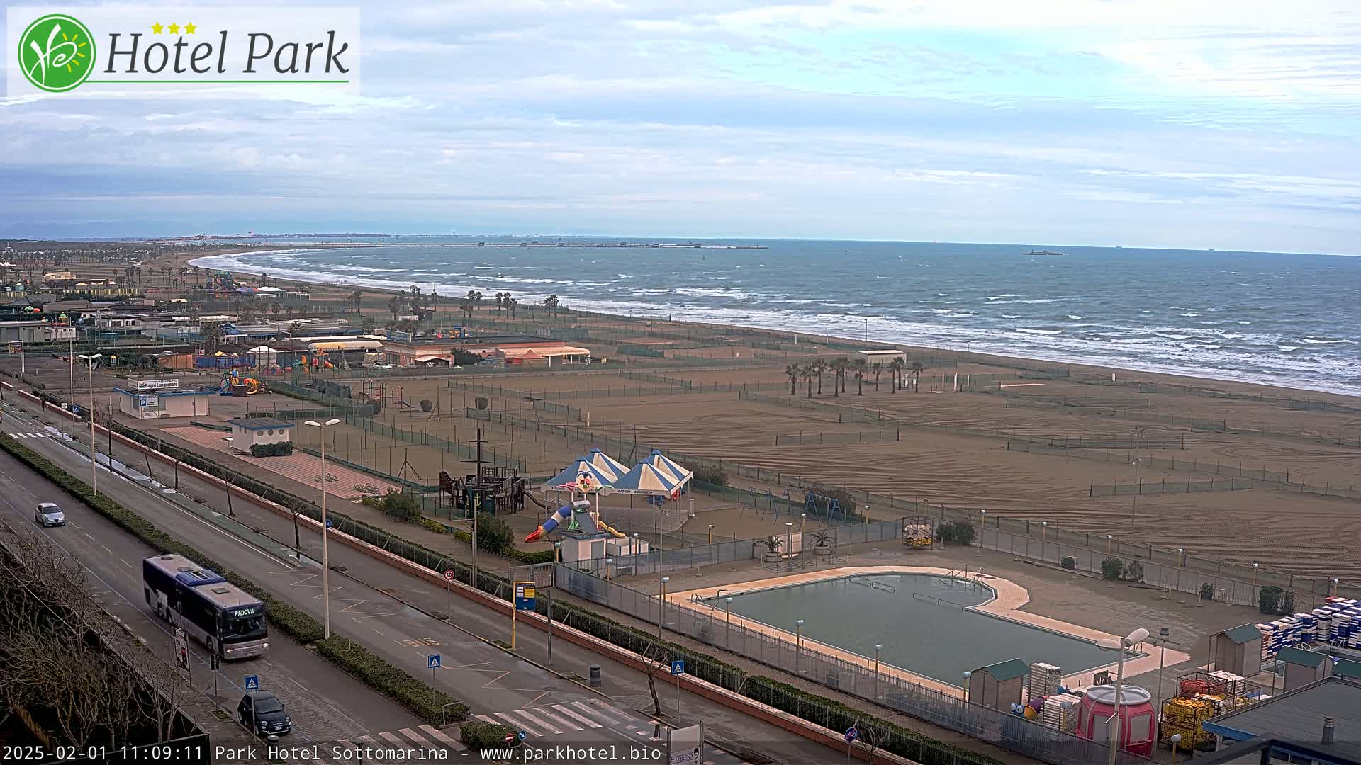 A beach with a playground, swimming pool, and roadway is visible, overlooking a wavy ocean under a partly cloudy sky.