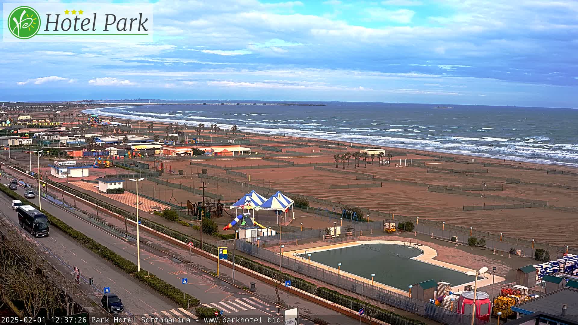 A beachside resort with a swimming pool, playground, and a sandy beach with wavy seas under a partly cloudy sky.