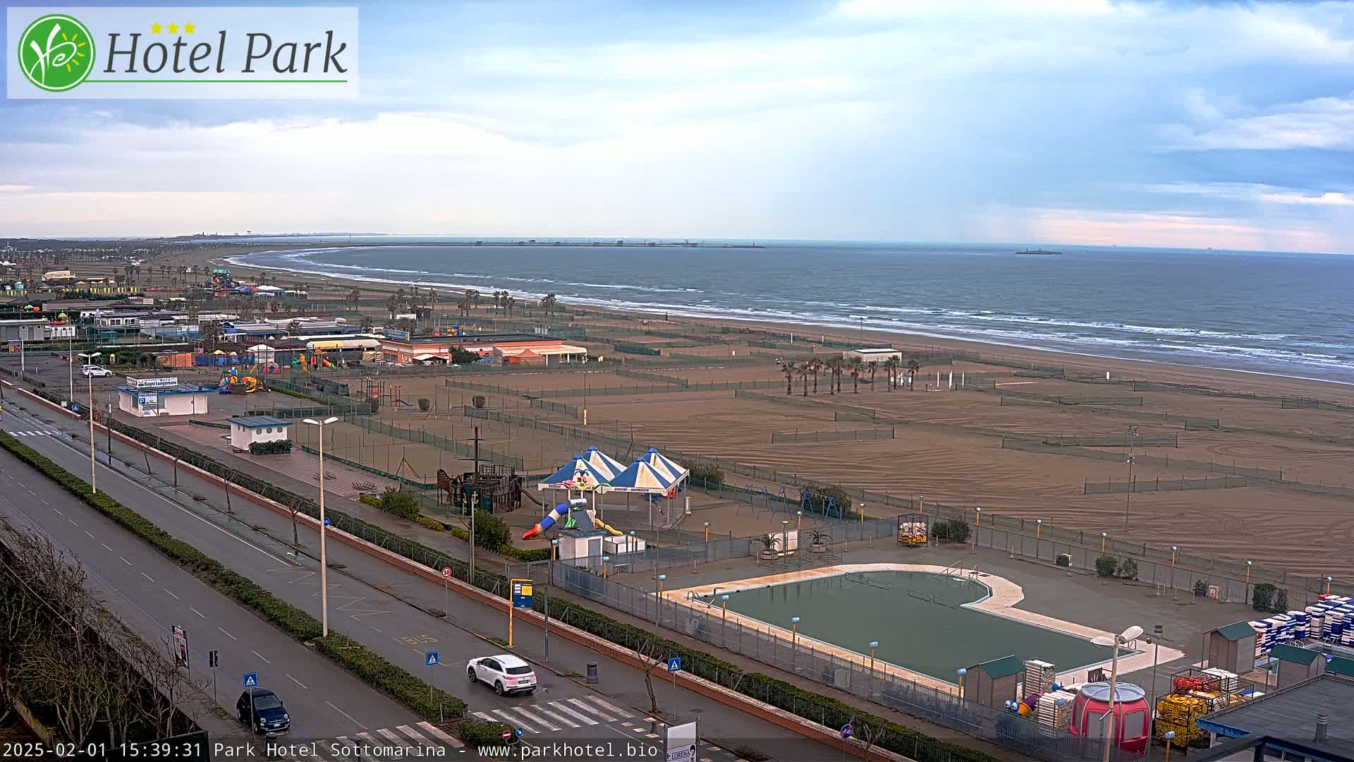 A beach with a playground, swimming pool, and a road is visible, overlooking a calm sea under a cloudy sky.