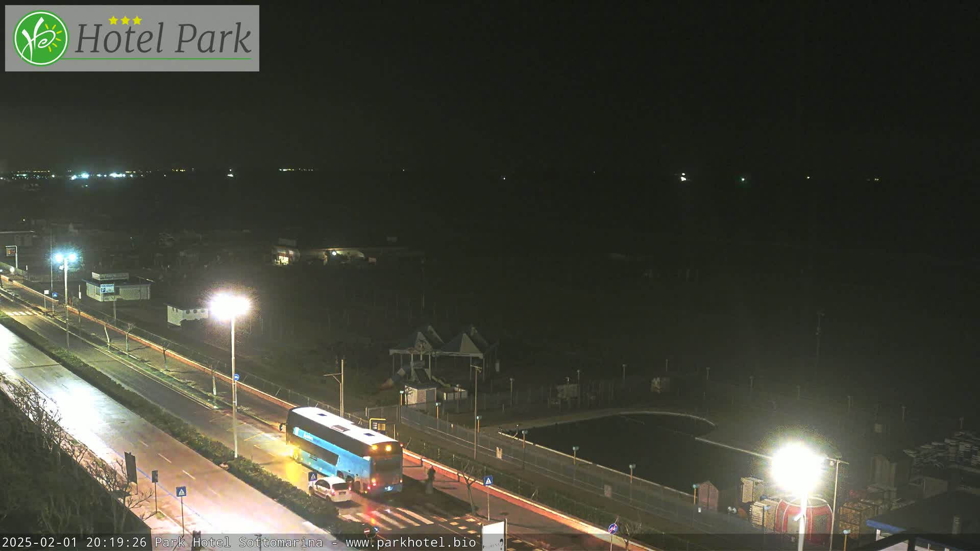 A nighttime, slightly overcast view shows a road with a bus and car, next to a dark body of water with a playground and pool visible across the road.
