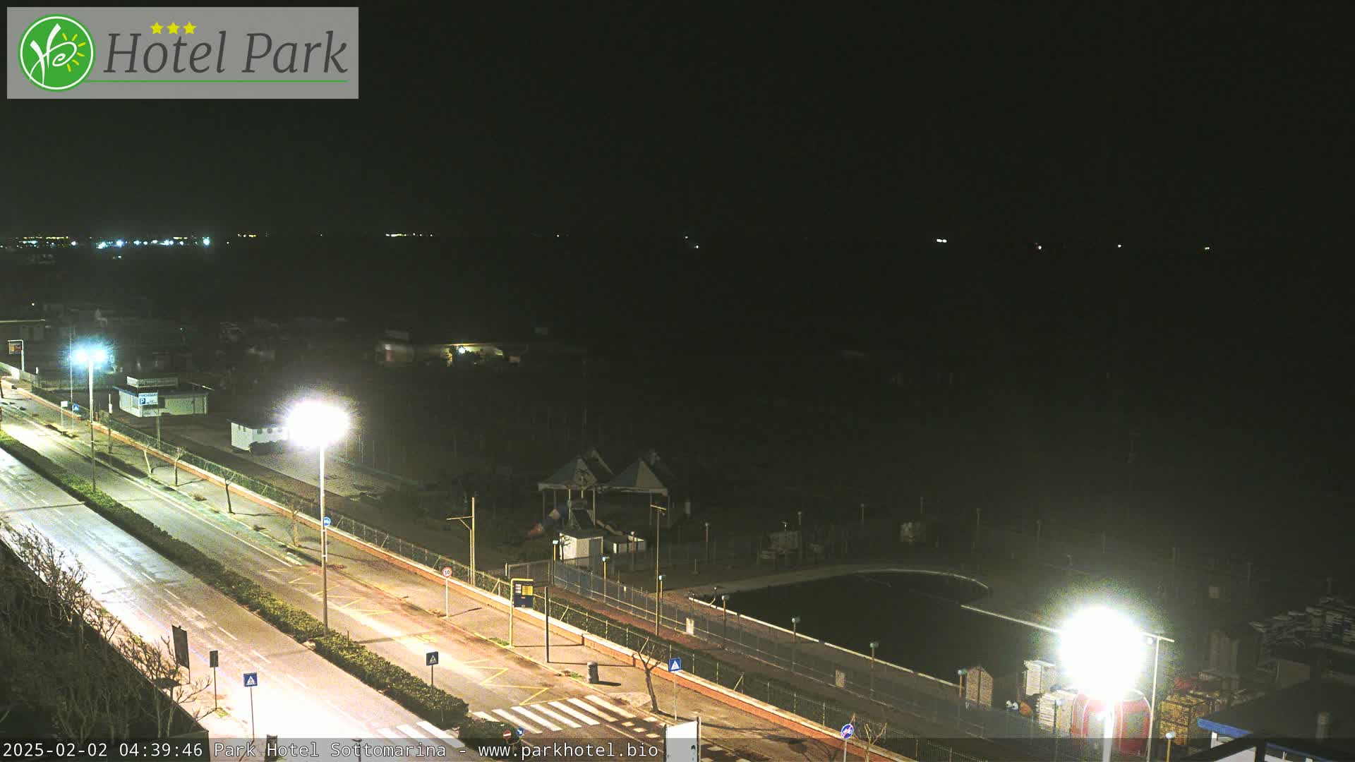 A nighttime view of a road next to a dark body of water, with lit parking areas, a playground, and a pool visible.
