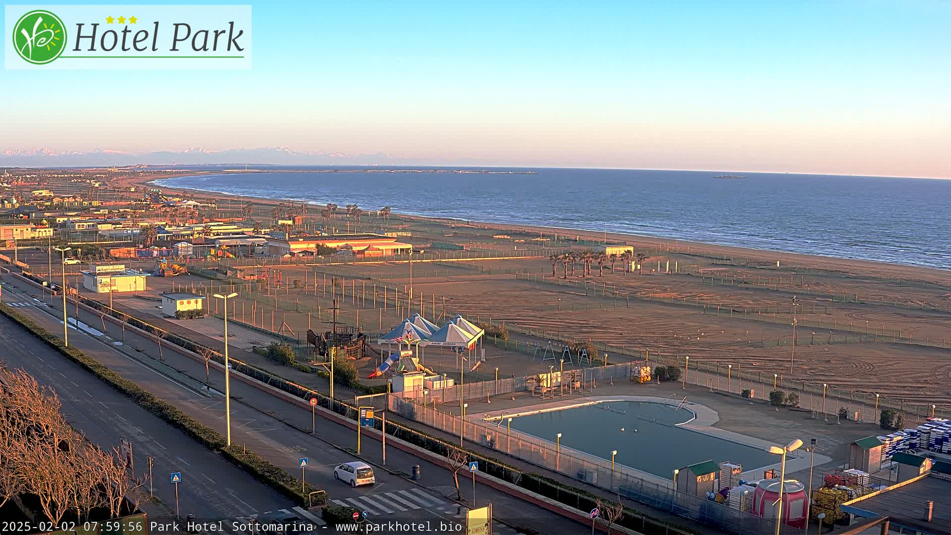 A sandy beach and calm ocean are visible under a clear, bright sky, with a playground, swimming pool, and road adjacent to the shore.