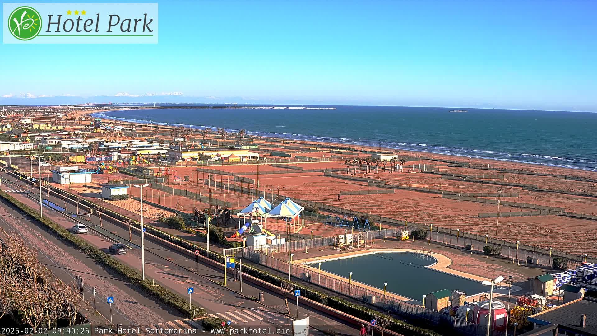 A sunny day at a beachfront resort shows a swimming pool, playground equipment, and a sandy beach with calm ocean waves in the background.