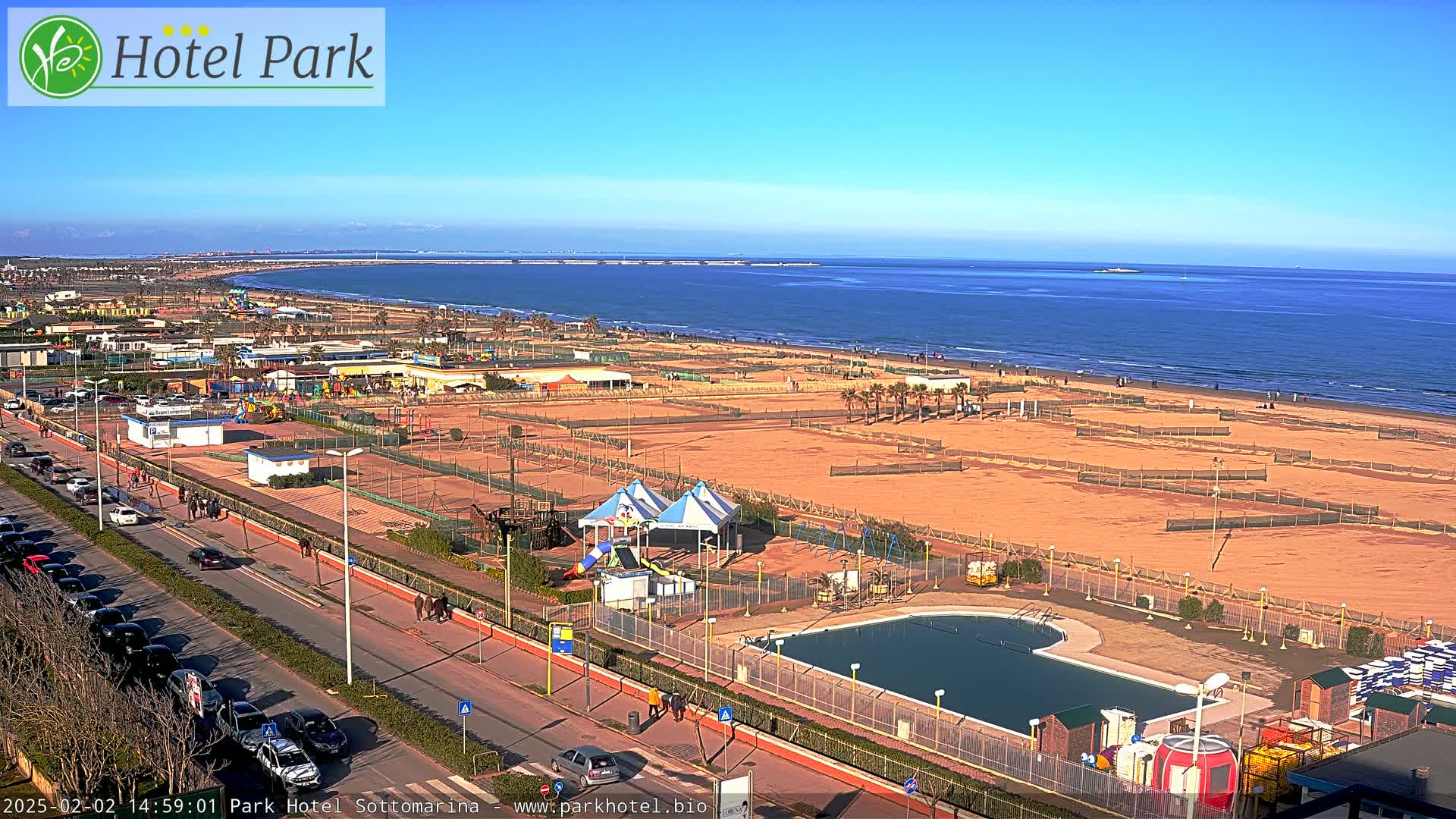 A seaside resort on a sunny day features a sandy beach, a swimming pool, playground equipment, and a road with parked cars.