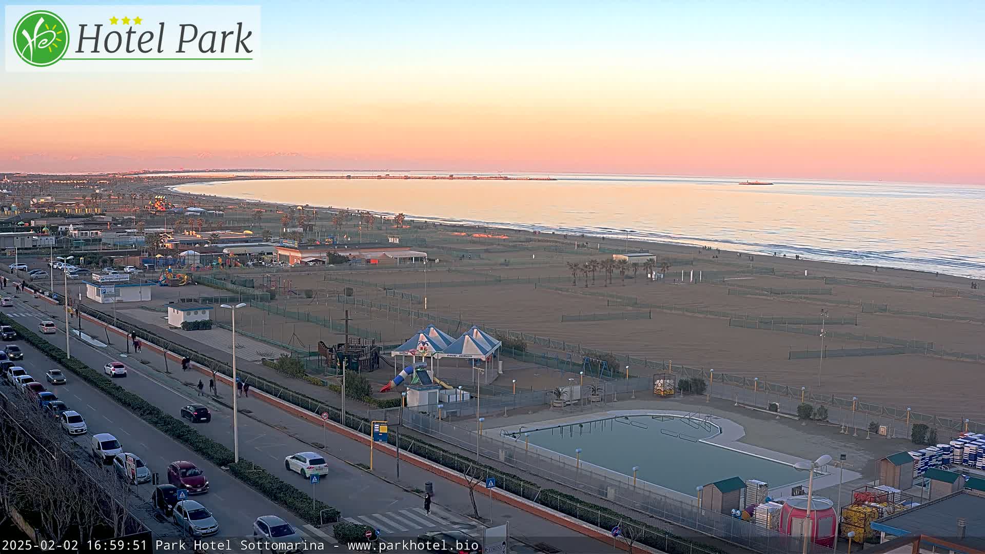 A beach with a playground, swimming pool, and road is visible at sunset, with calm ocean waters in the background.
