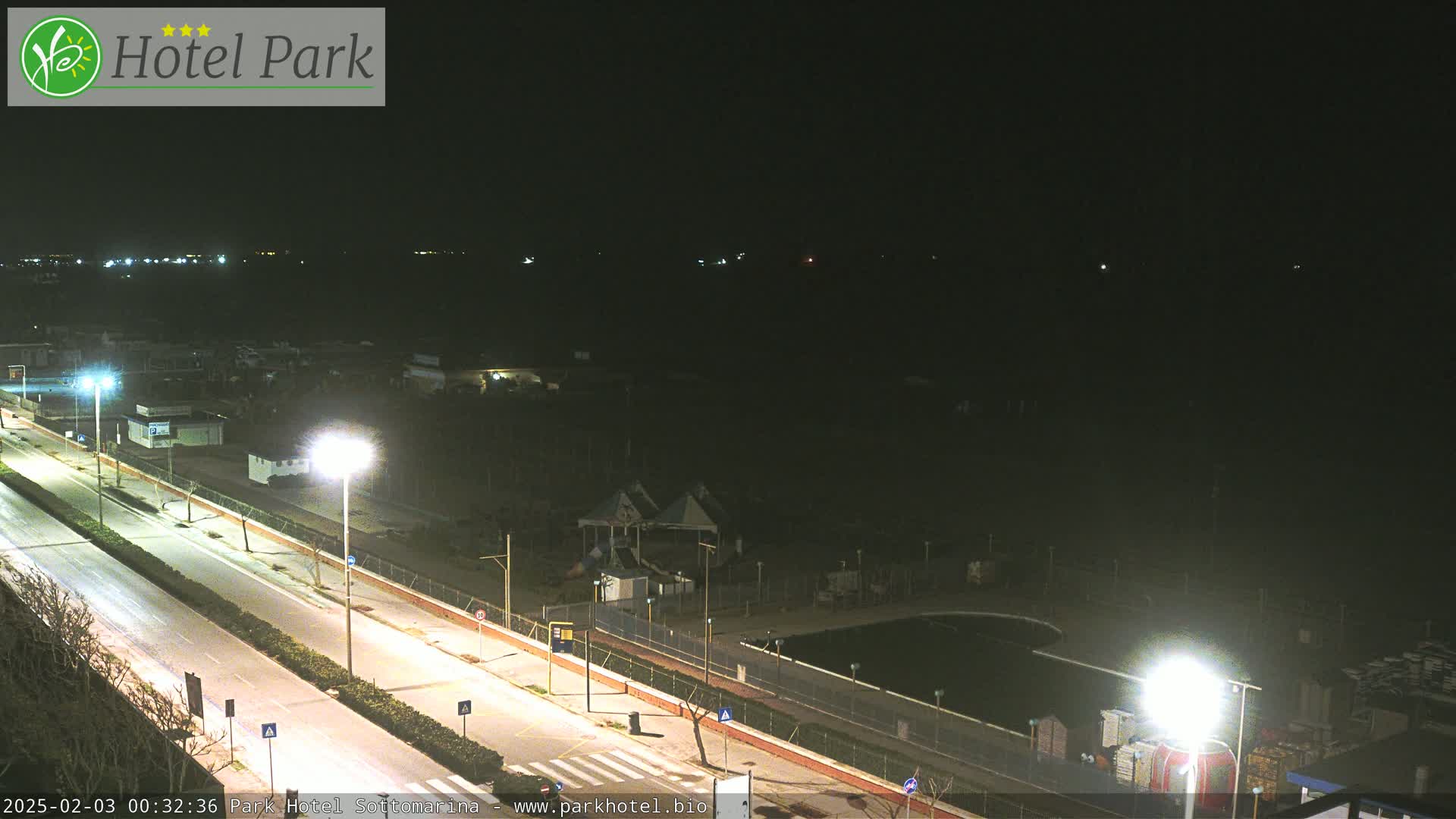 A nighttime view of a mostly empty road next to a seaside park with a pool and playground, under a dark, clear sky.
