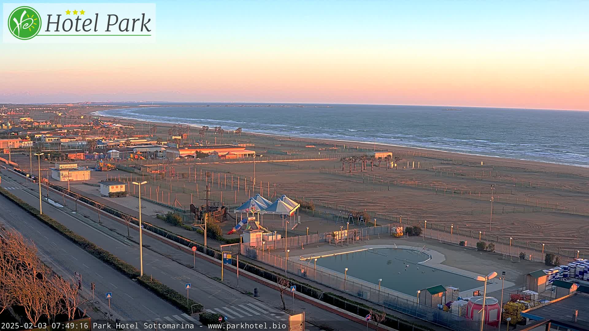 A beachside view at sunset shows a sandy beach, calm ocean waves, a swimming pool, playground equipment, and a roadway leading to buildings.