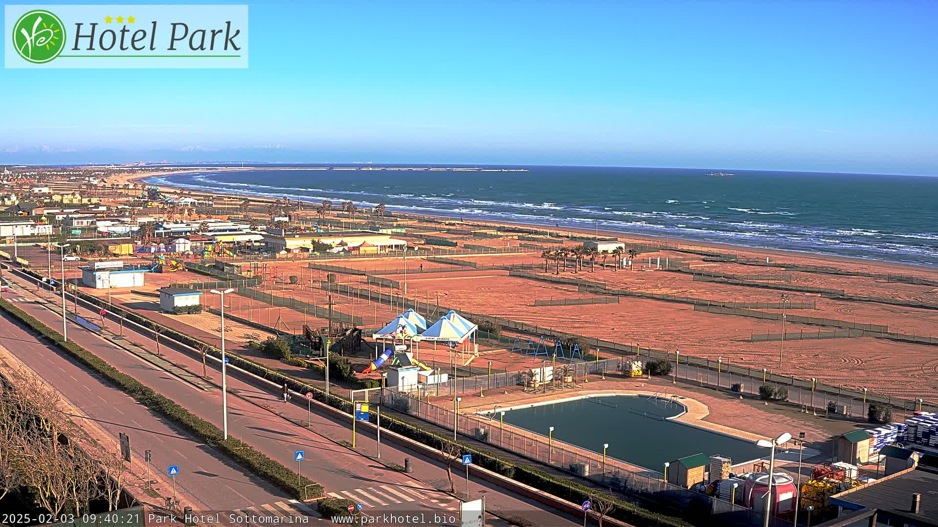 An aerial view shows a beach with a wavy ocean under a clear blue sky, next to a coastal road and recreational area with a swimming pool and playground equipment.